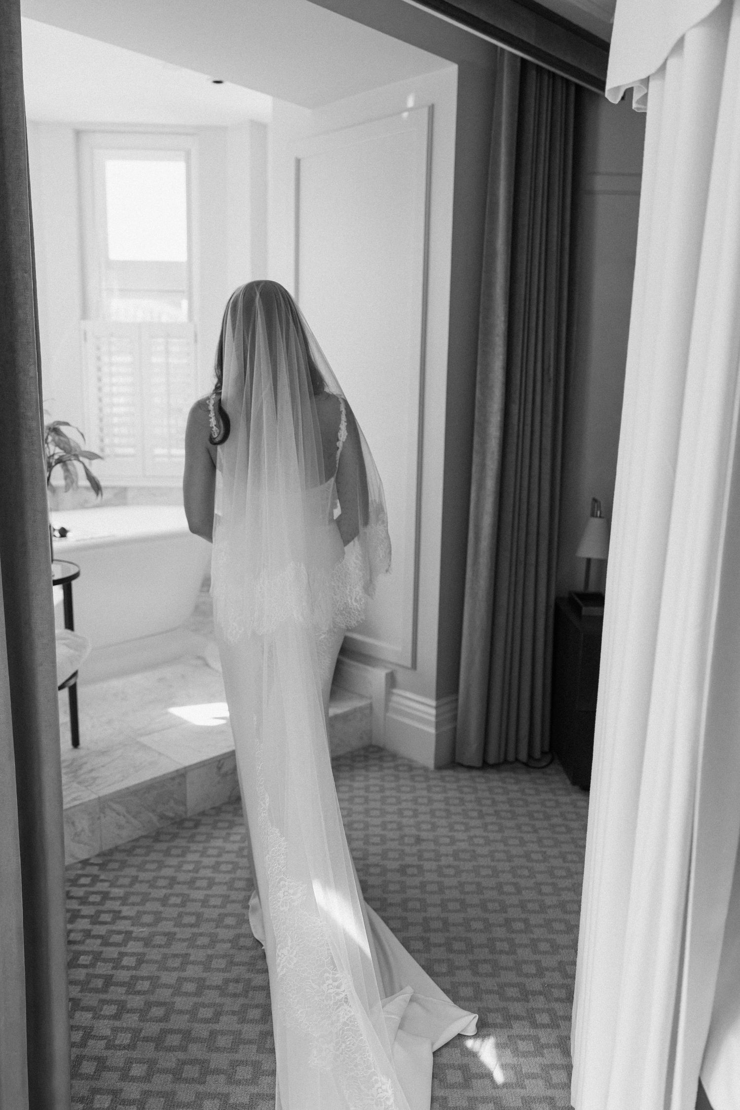 Black and white image of a bride in a fitted wedding gown with cathedral veil walking through a hotel room doorway.