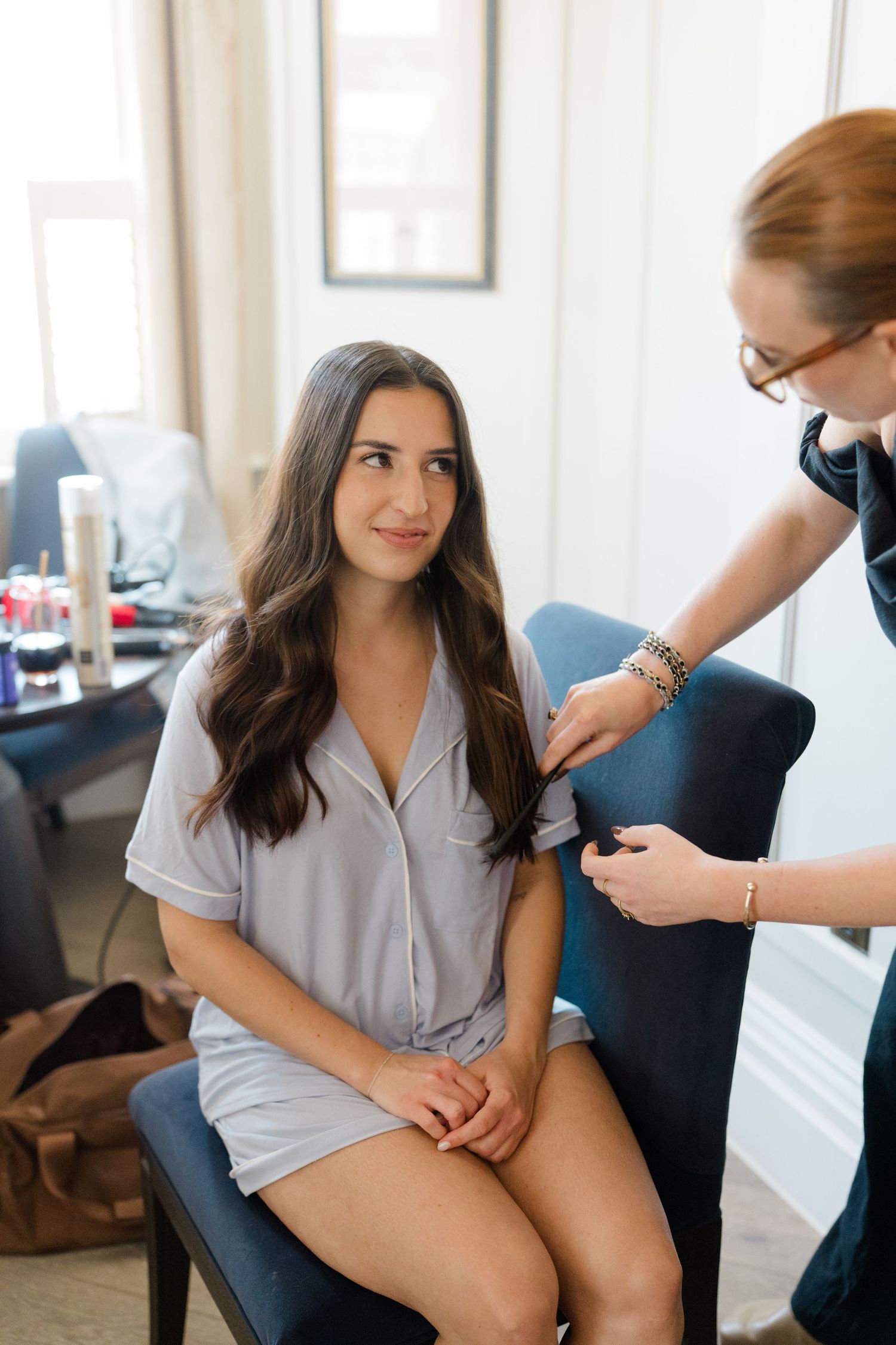 Bride getting her long dark hair styled by a redheaded hairstylist in a bright hotel room before her wedding.