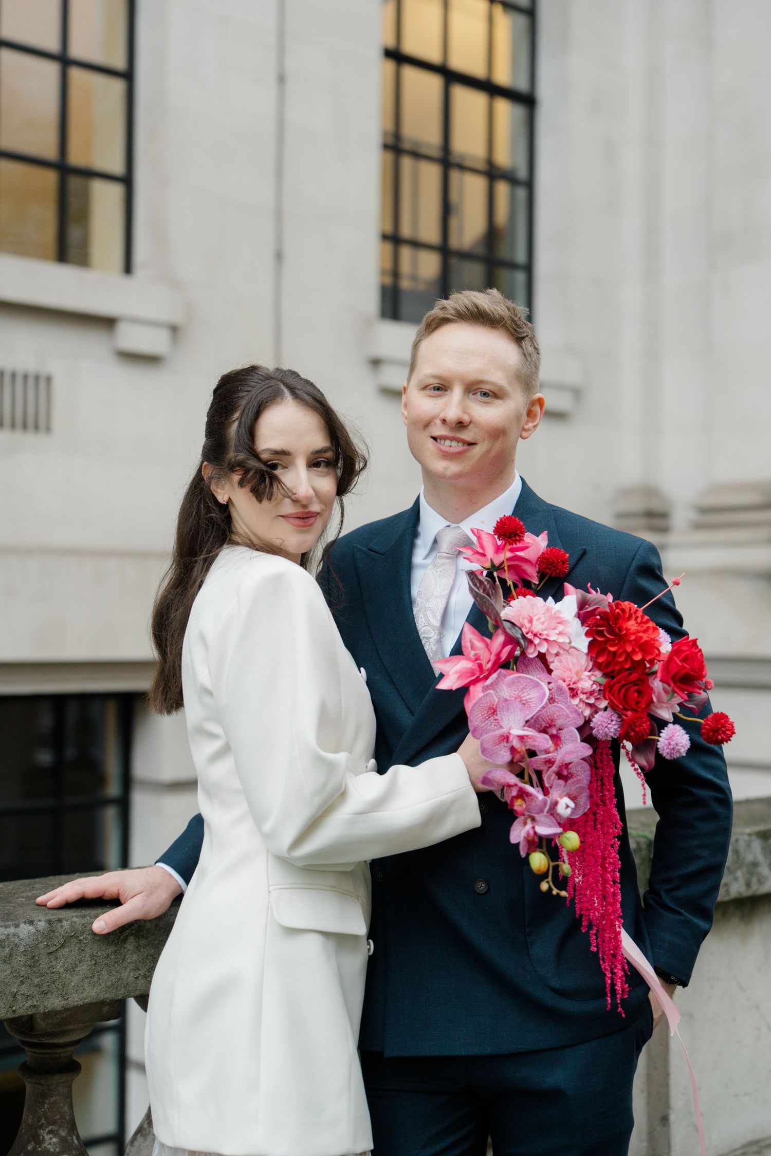 Elegant couple poses with vibrant pink and red bridal bouquet outside a classic building on their wedding day.