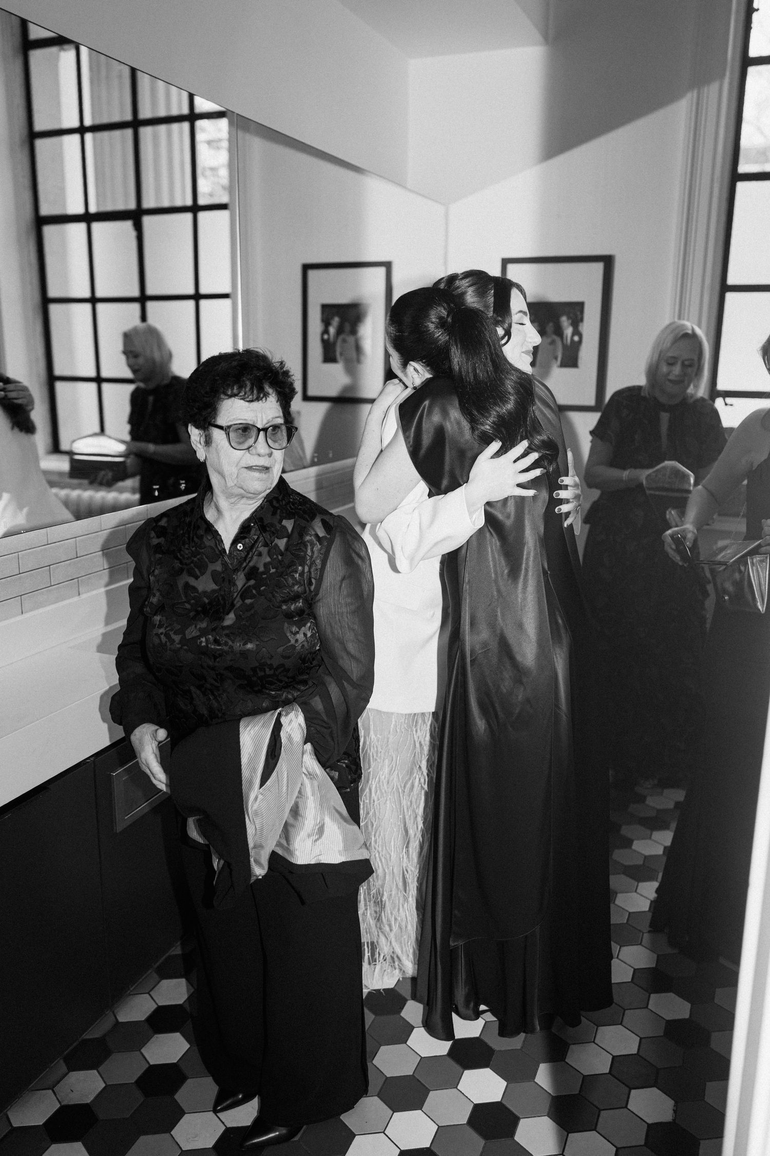 Black and white photo of bride hugging guest while elderly woman stands nearby at wedding venue.