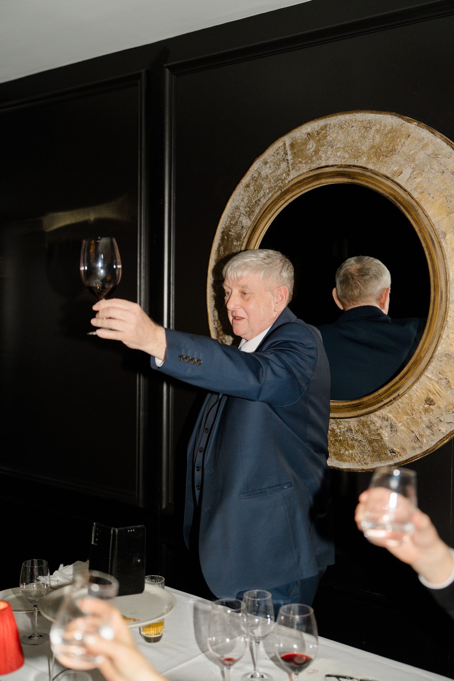 Older gentleman in navy suit raises glass of red wine for toast at elegant wedding reception.