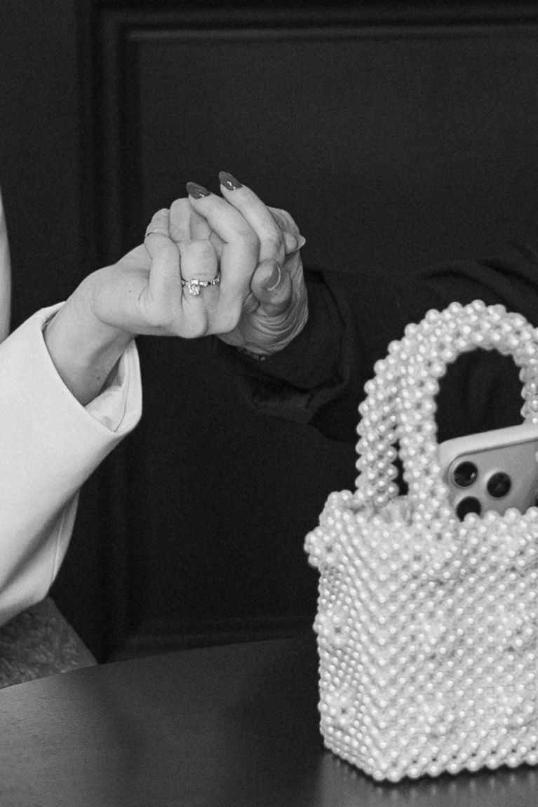 Black and white photo of two hands holding, wearing rings, with a pearl beaded handbag on a table.
