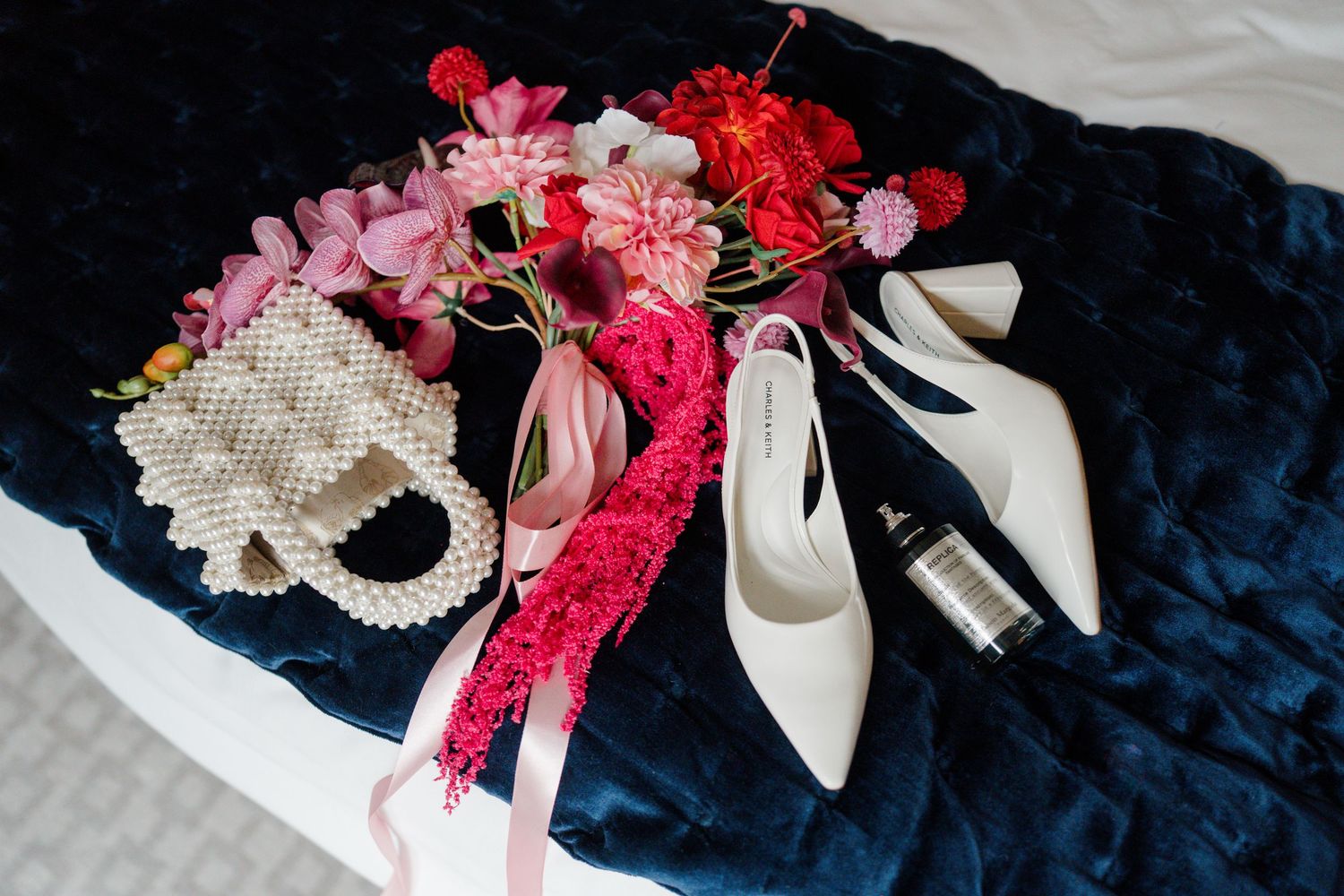 White heels, pearl clutch, and vibrant pink and red floral bouquet arranged on a navy velvet surface.