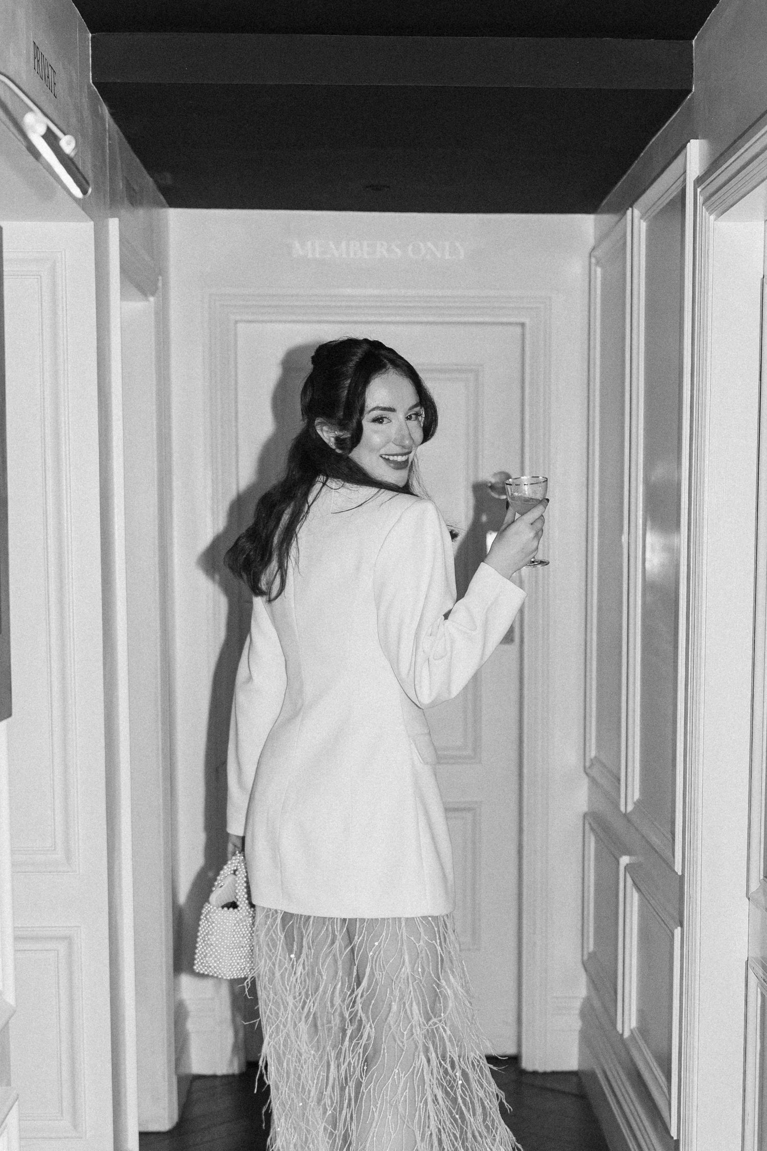 Bride in white feather dress holding cocktail at members only door, black and white.