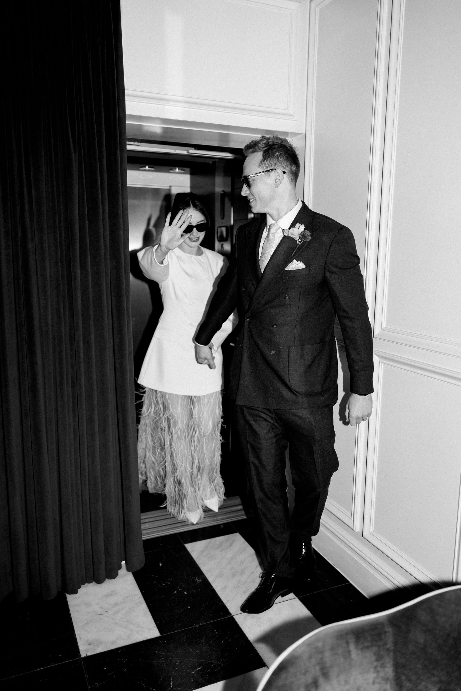 Black and white photo of newlyweds exiting elevator, bride waving in white mini dress with feathers.
