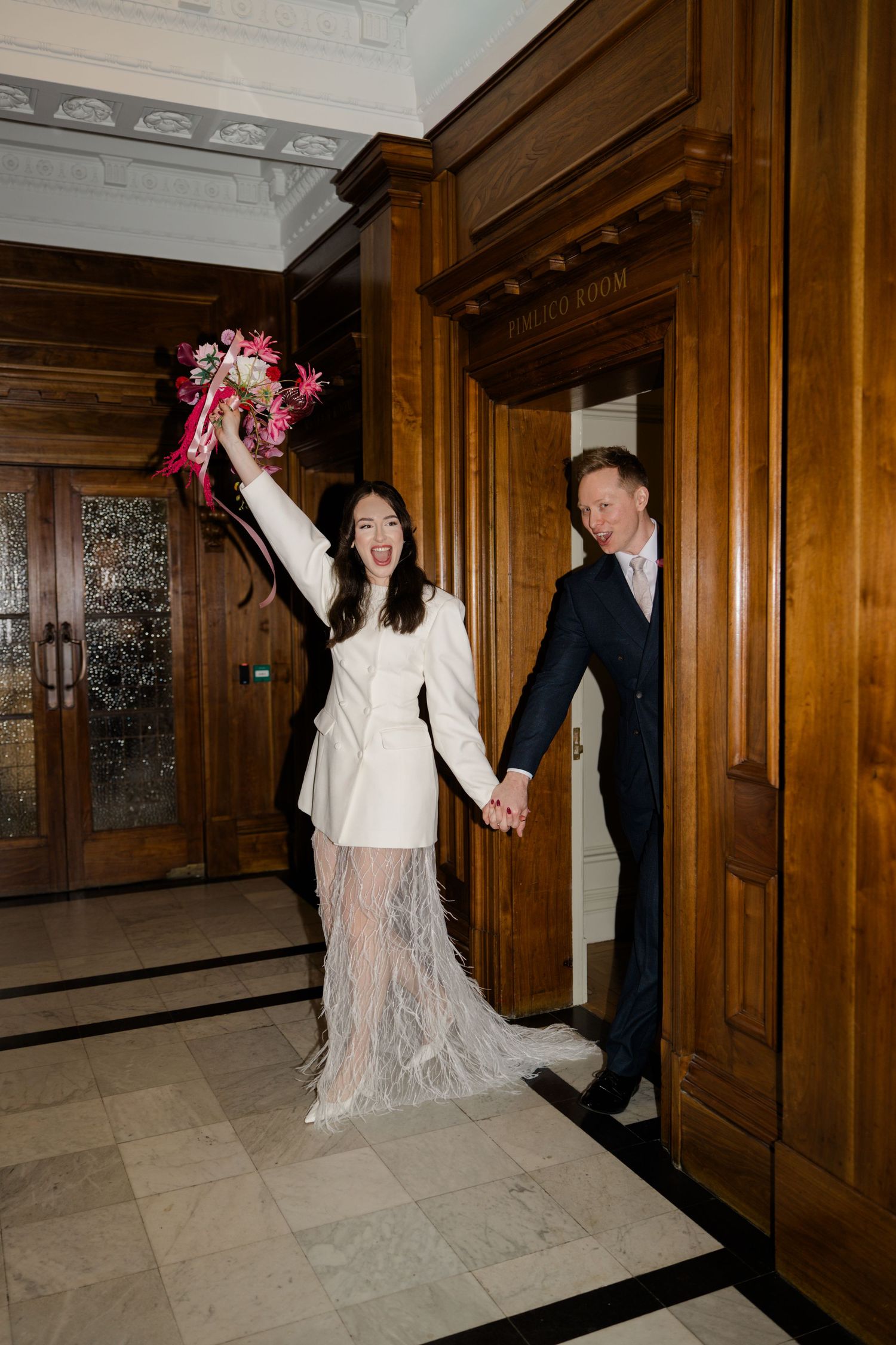 Bride in white blazer feather skirt raises bouquet exiting registry office with groom.