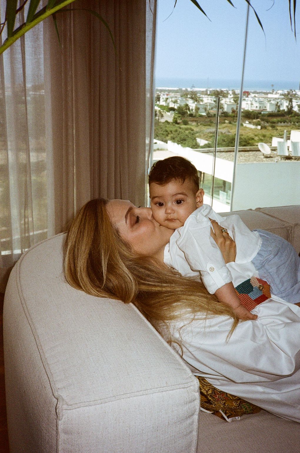 A woman with blonde hair kisses a baby in white clothing while relaxing on a cream sofa near a large window.