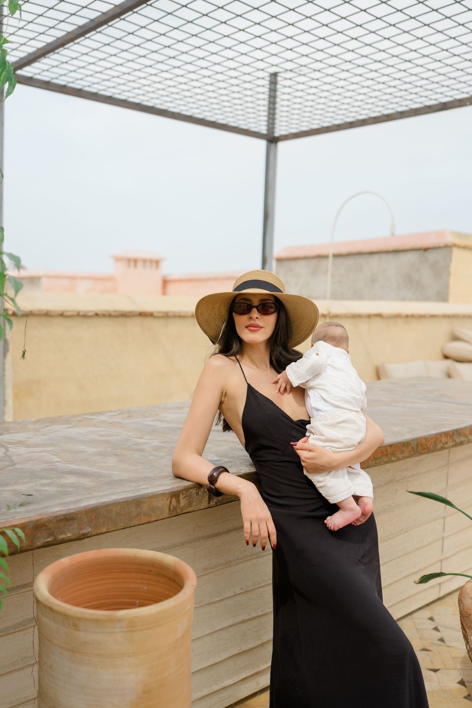 Stylish woman in black dress and sun hat holding a baby on a rooftop terrace.