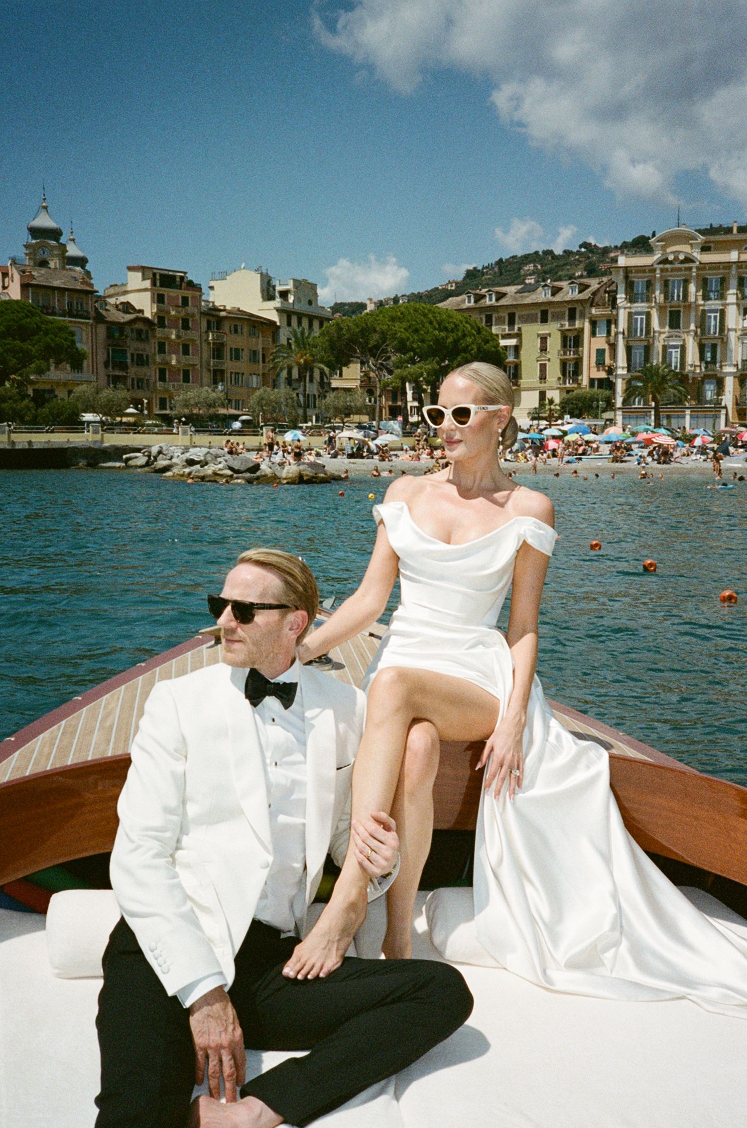 Elegant couple in wedding attire on a wooden boat in Italian Riviera, bride in white gown with sunglasses, groom in white tuxedo.