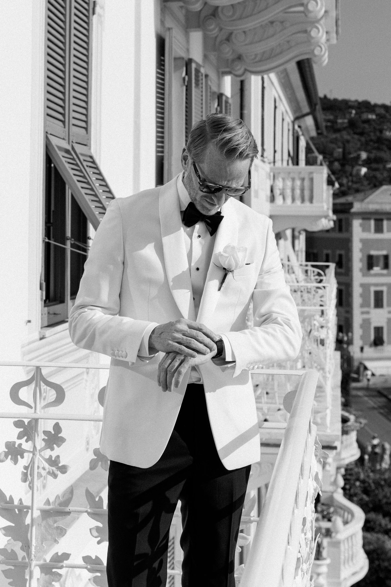 Dapper groom in white tuxedo jacket adjusts cufflinks on ornate hotel balcony before wedding.