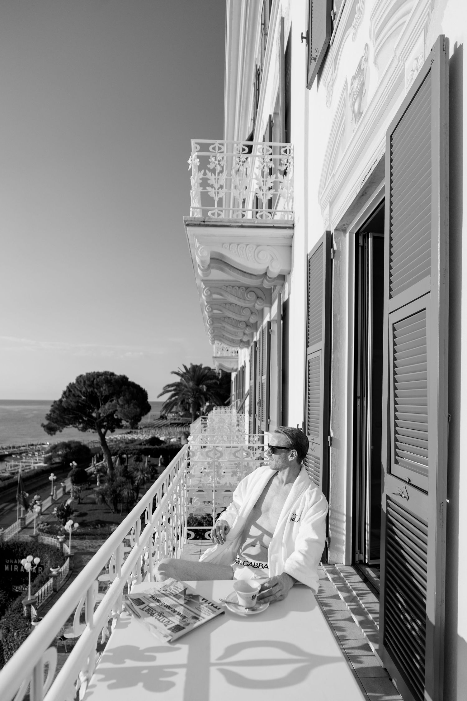 Man in white robe reads magazine on ornate hotel balcony overlooking Mediterranean coastline.