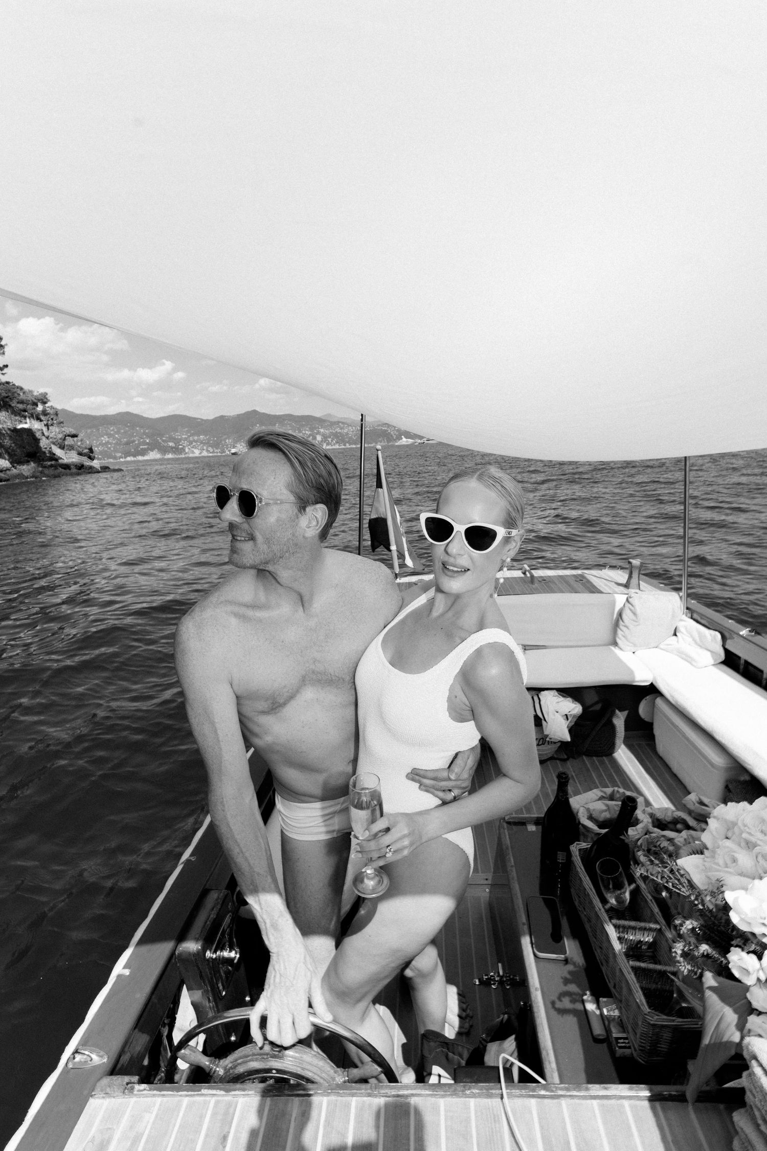 Couple in swimwear steering a classic wooden boat along scenic Italian Mediterranean coastline.