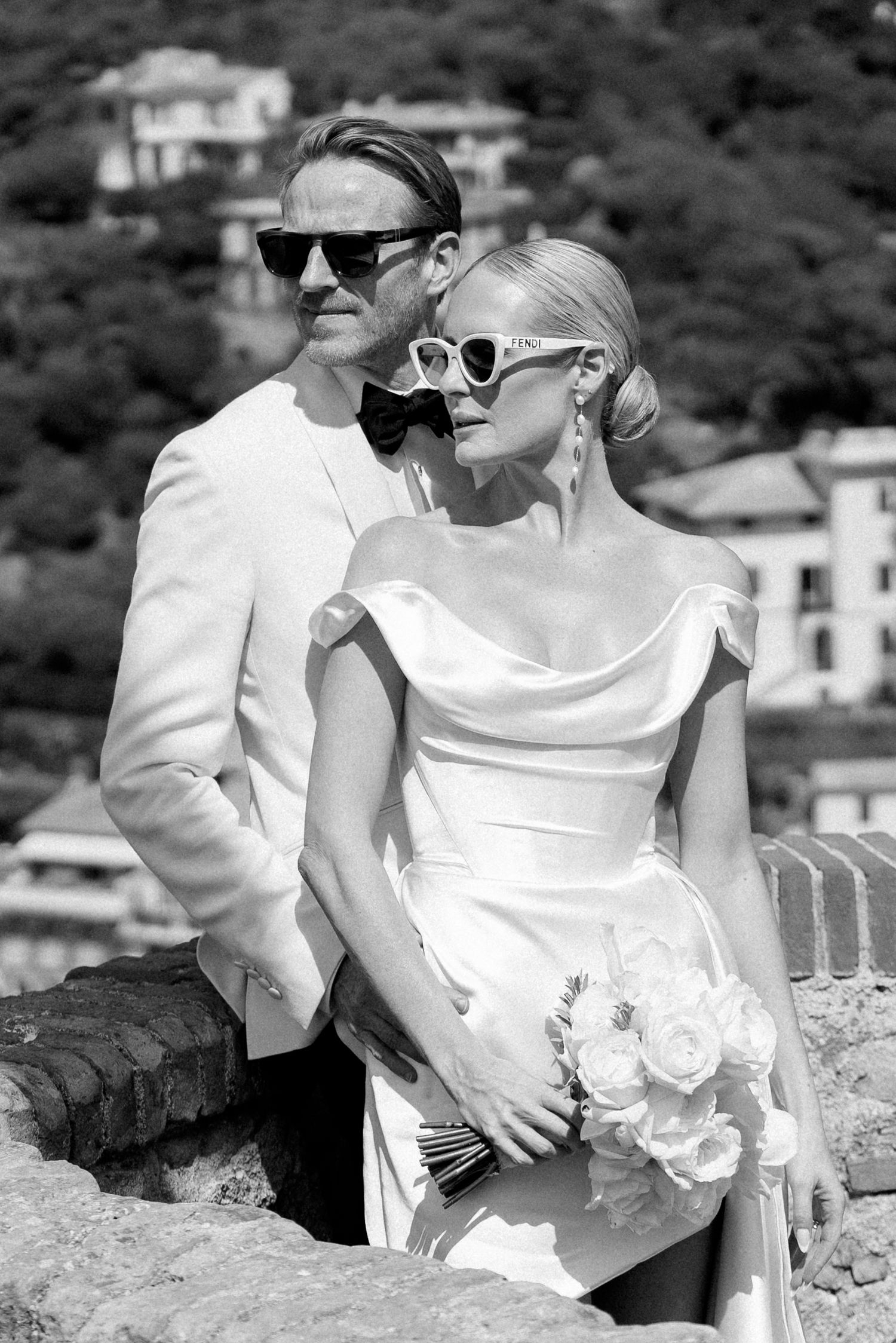 Stylish newlyweds in sunglasses pose against stone wall with Italian hillside backdrop, bride holding white roses.