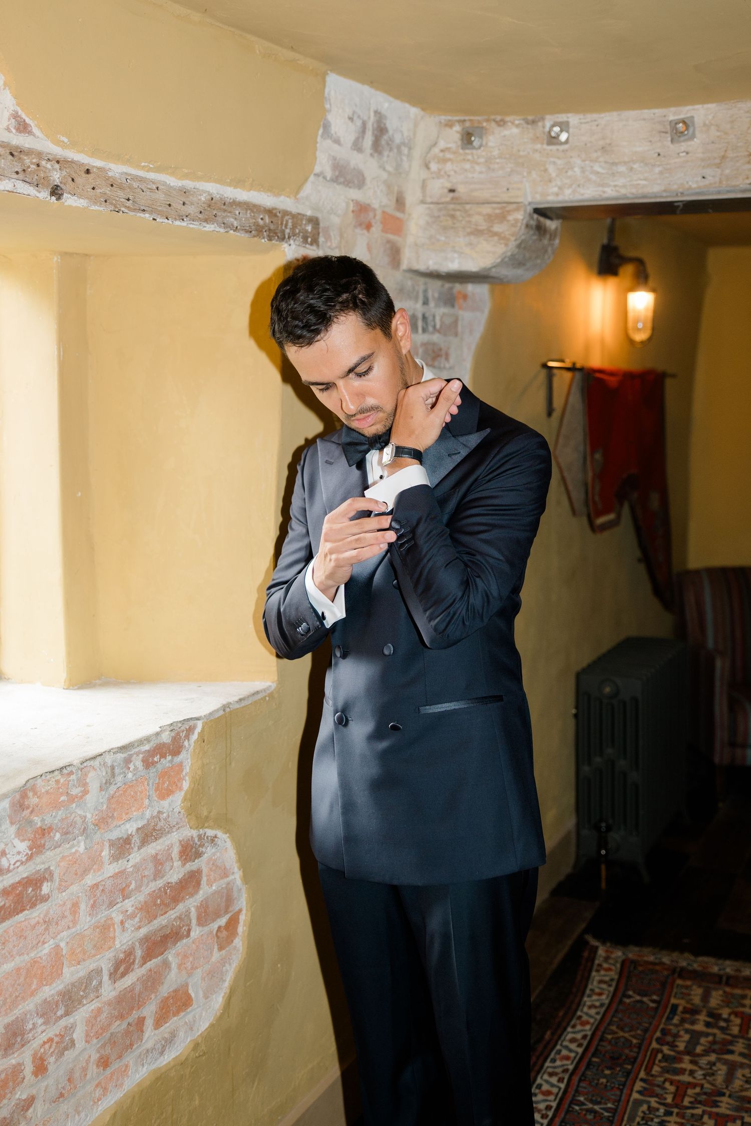 Elegant groom in navy tuxedo adjusting cufflinks in rustic venue with brick walls and warm lighting.