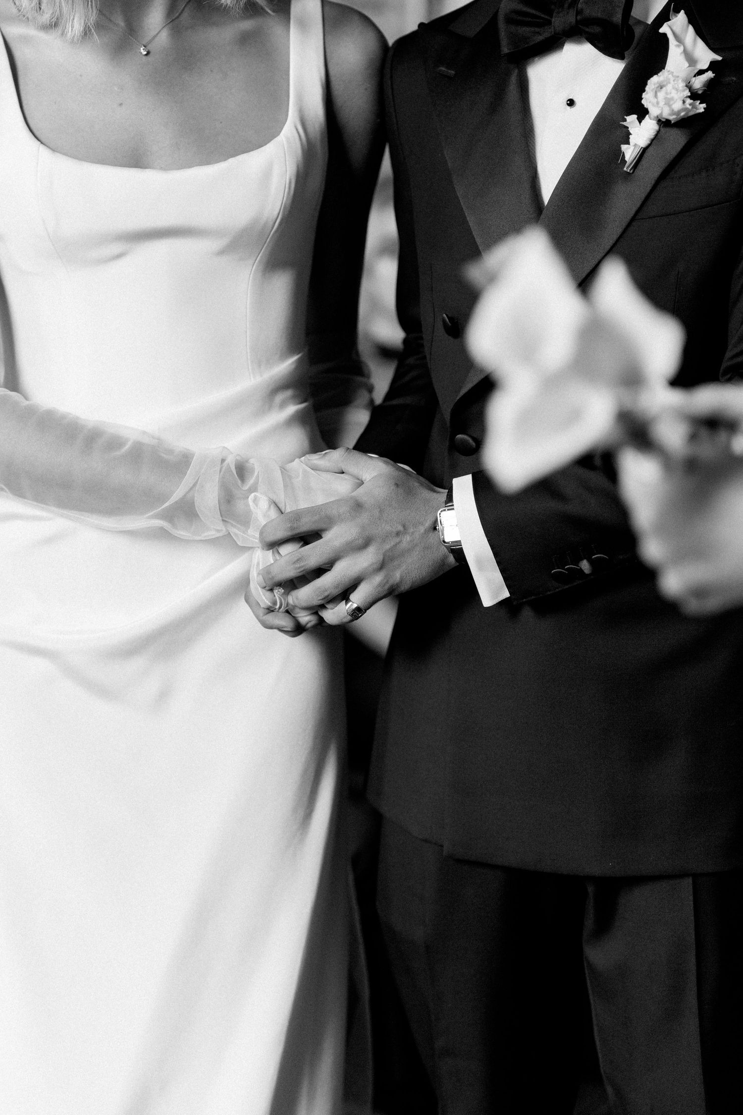 Black and white close-up of bride and groom holding hands, bride wearing white gloves and groom in a tuxedo.