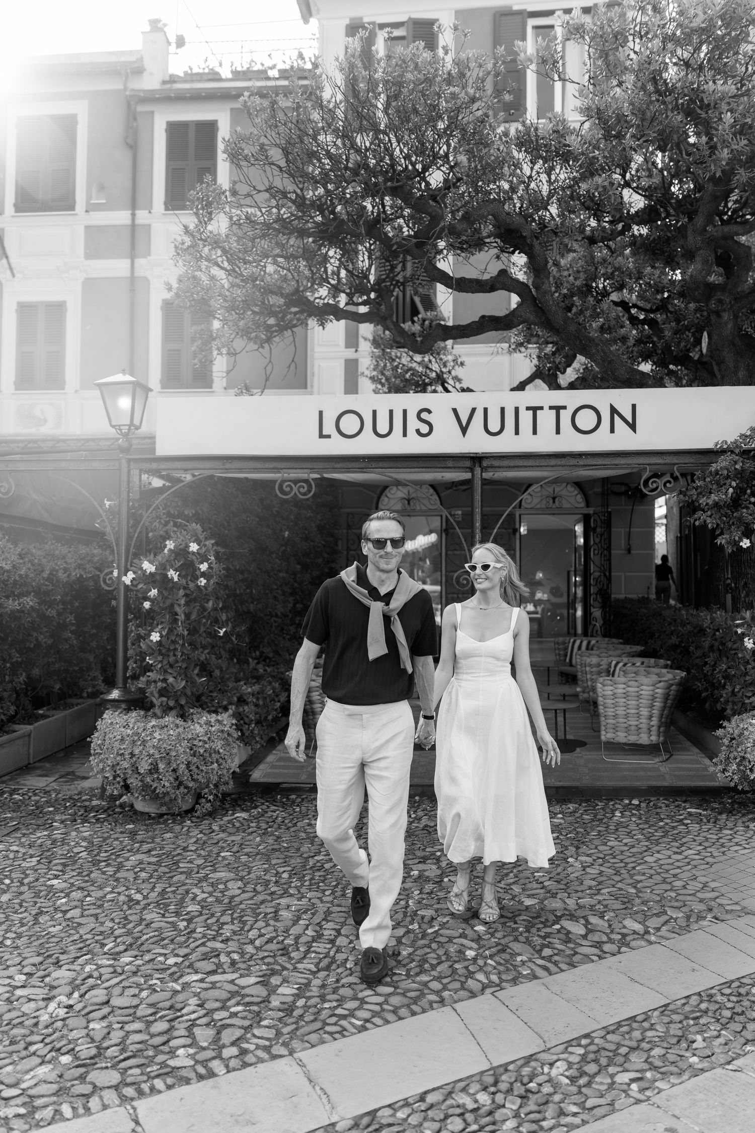 Stylish couple holding hands walking past Louis Vuitton store on cobblestone street, black and white photo.