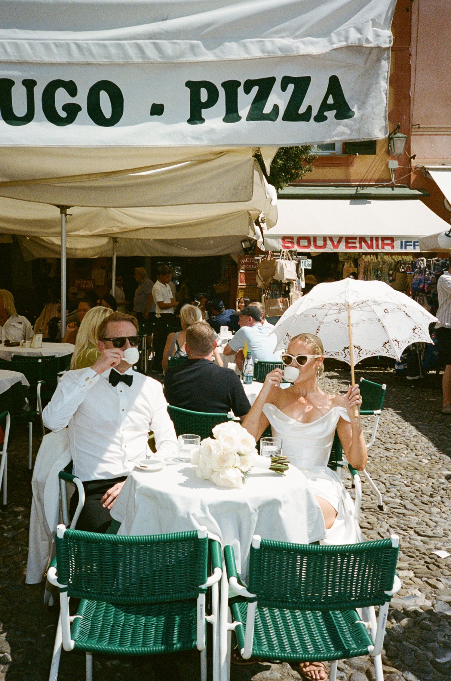 Newlyweds in wedding attire sipping coffee at an outdoor cafe in Portofino, Italy, with a white parasol nearby.