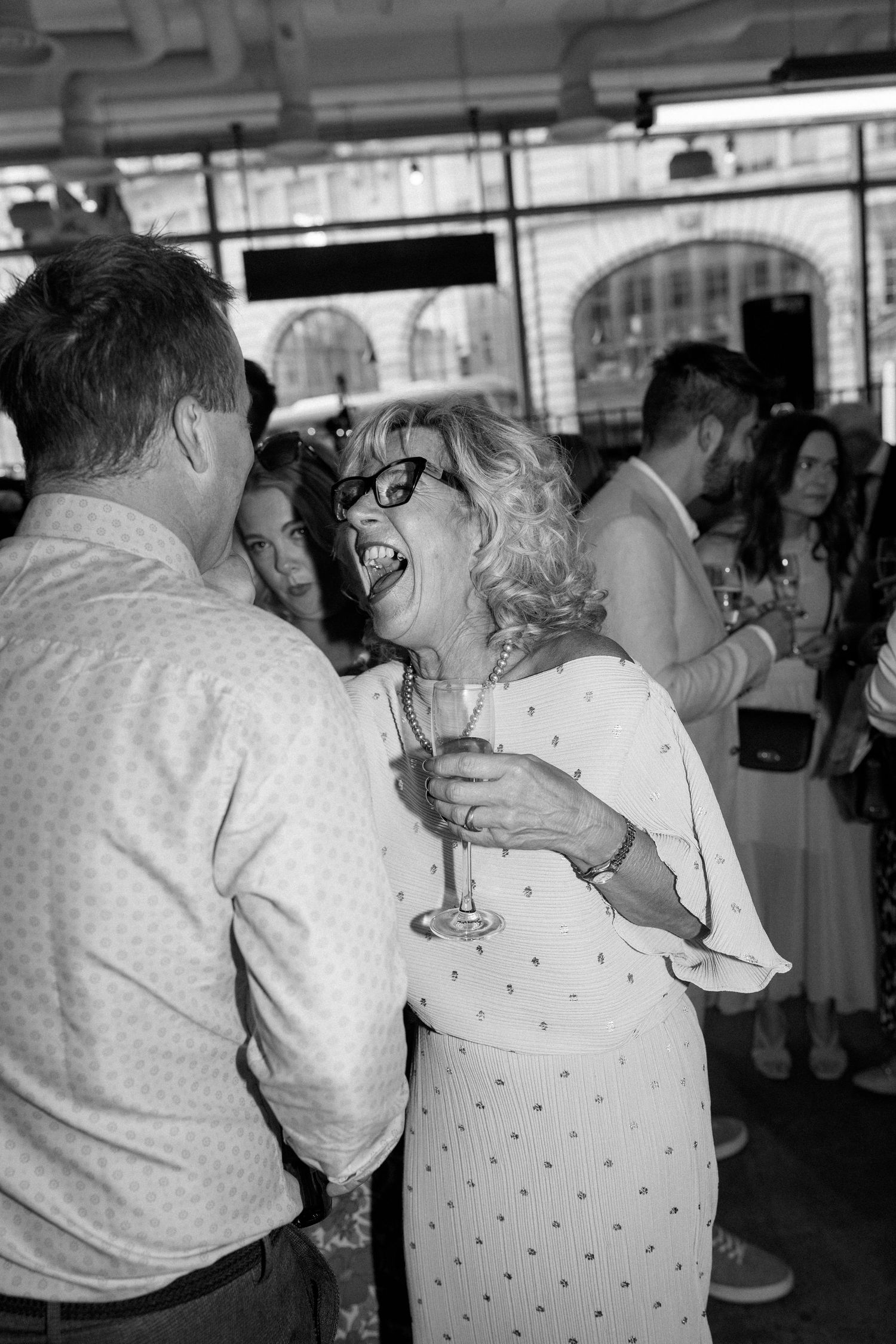 Woman in glasses laughing joyfully with champagne flute at lively wedding reception in black and white photo.
