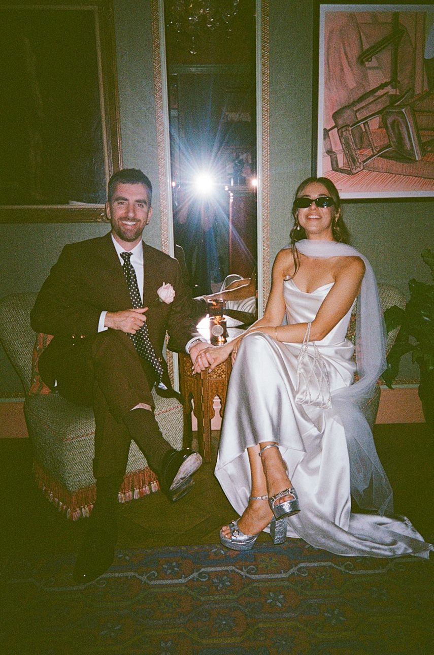 Bride in white satin gown with sunglasses and groom in dark suit sit together in warmly lit elegant room.