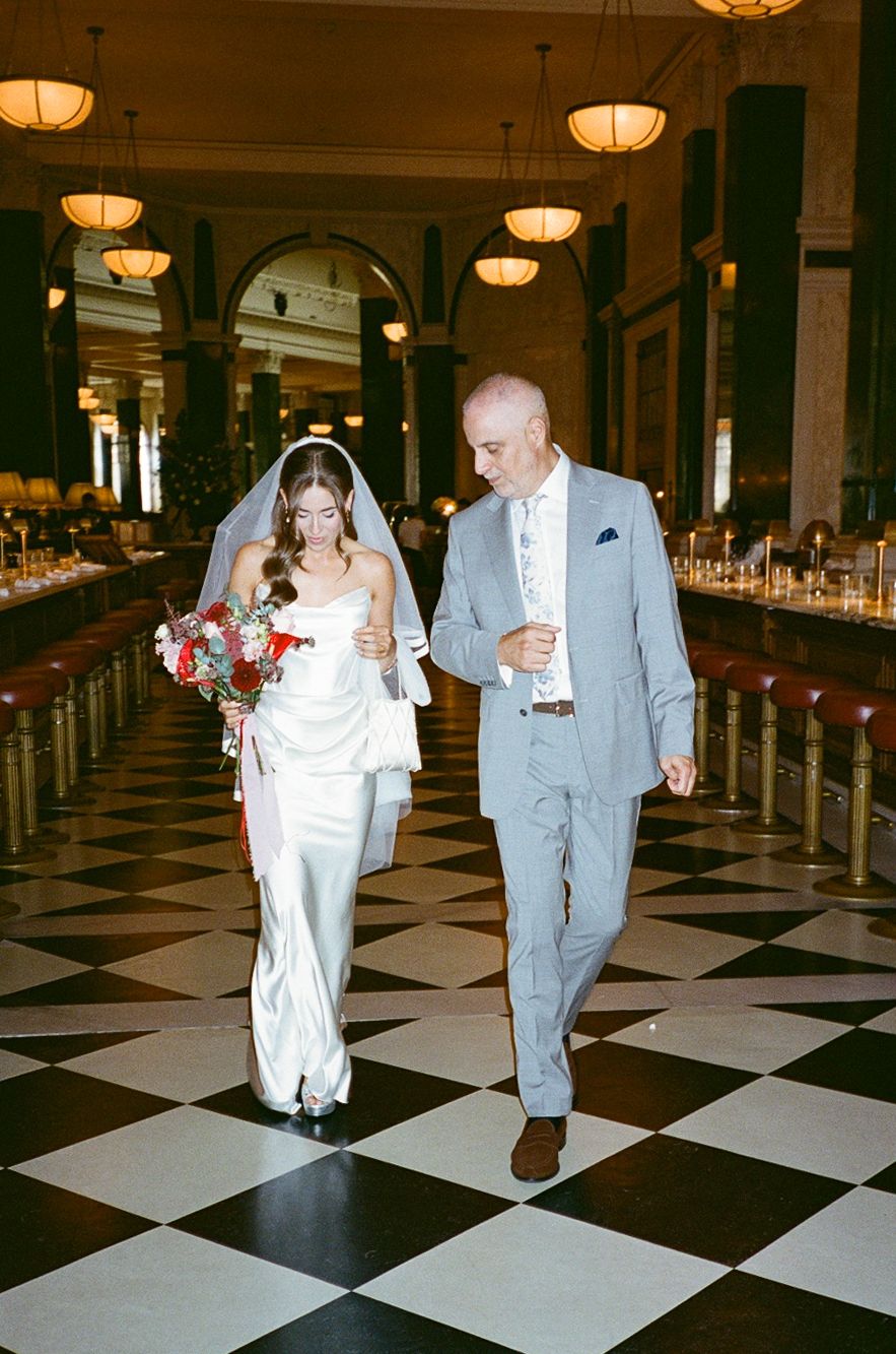 Bride in white gown with veil and red bouquet walks with suited man on checkered floor in elegant venue.