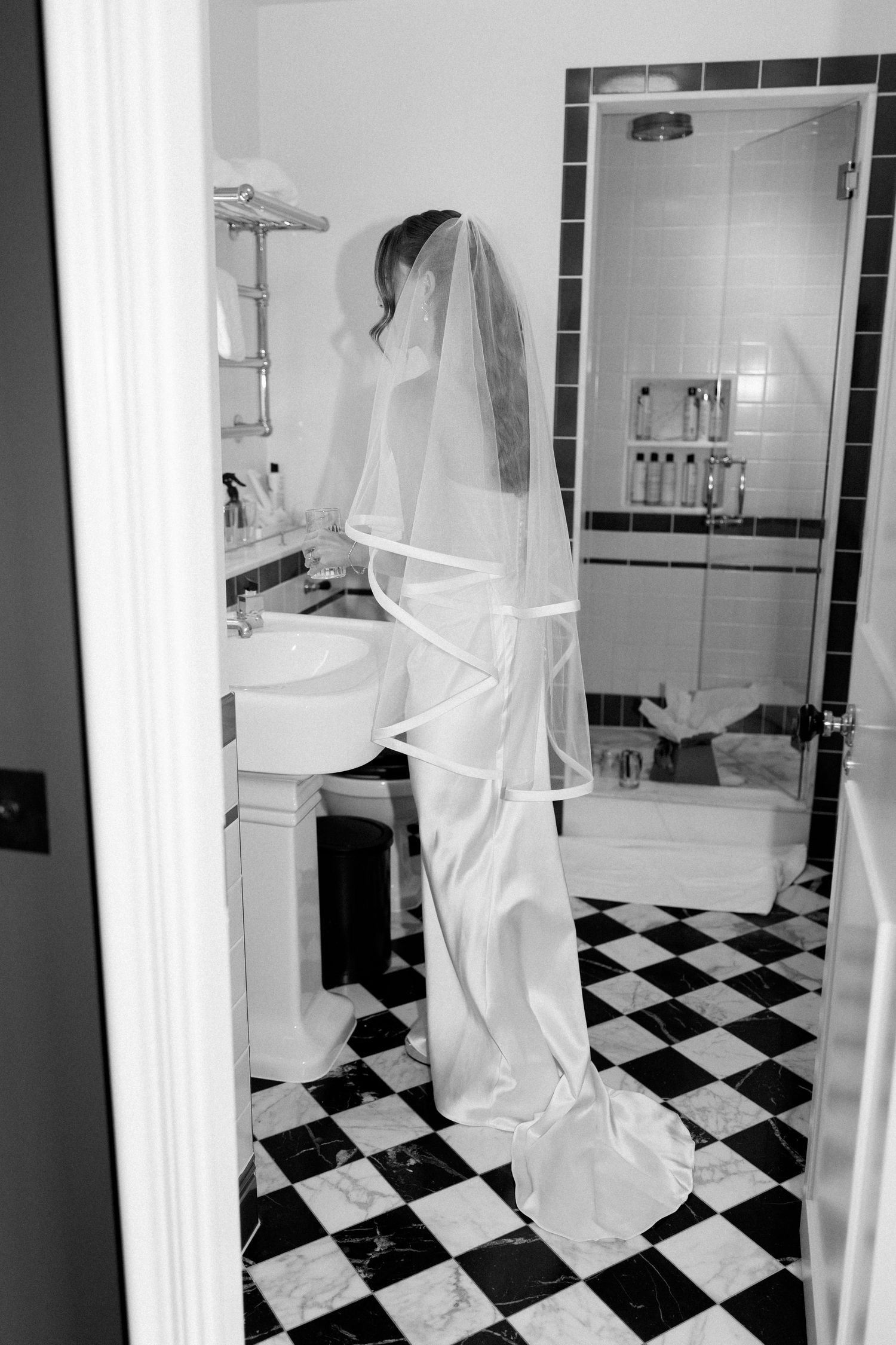 Black and white image of bride in wedding dress and veil standing in classic checkered bathroom.