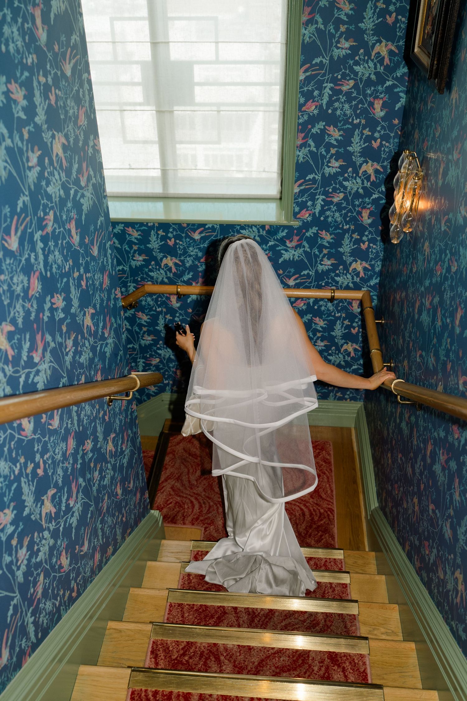 Bride in white gown and layered veil ascending red-carpeted staircase in room with ornate blue floral wallpaper.