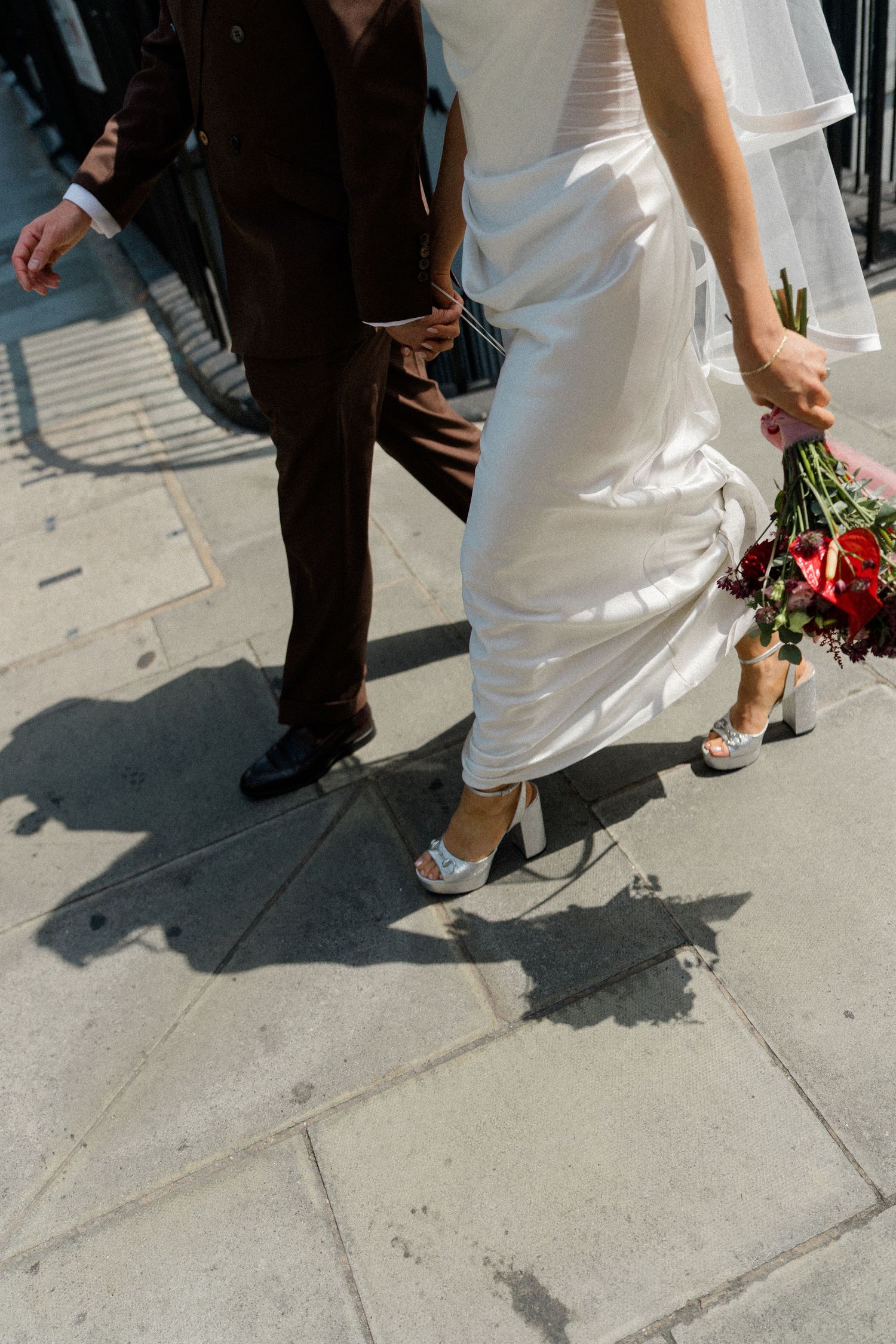 Close-up of bride in white satin dress and heels walking with groom on sunlit pavement holding bouquet.