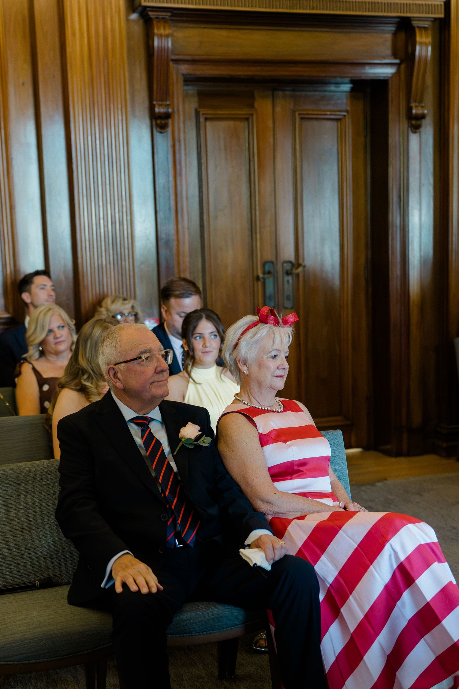 Wedding guests in formal attire seated in wooden paneled ceremony room, woman in red striped dress.