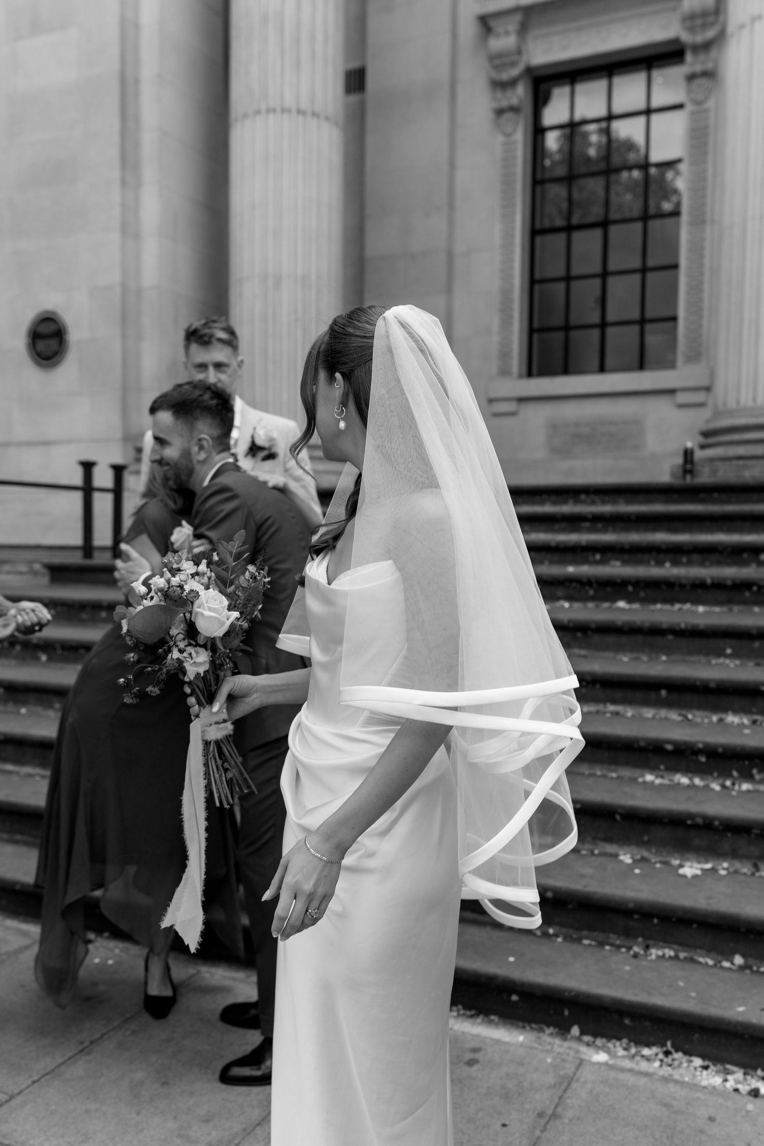 Bride in white gown and veil holds bouquet outside grand stone building as groom greets guests on steps.