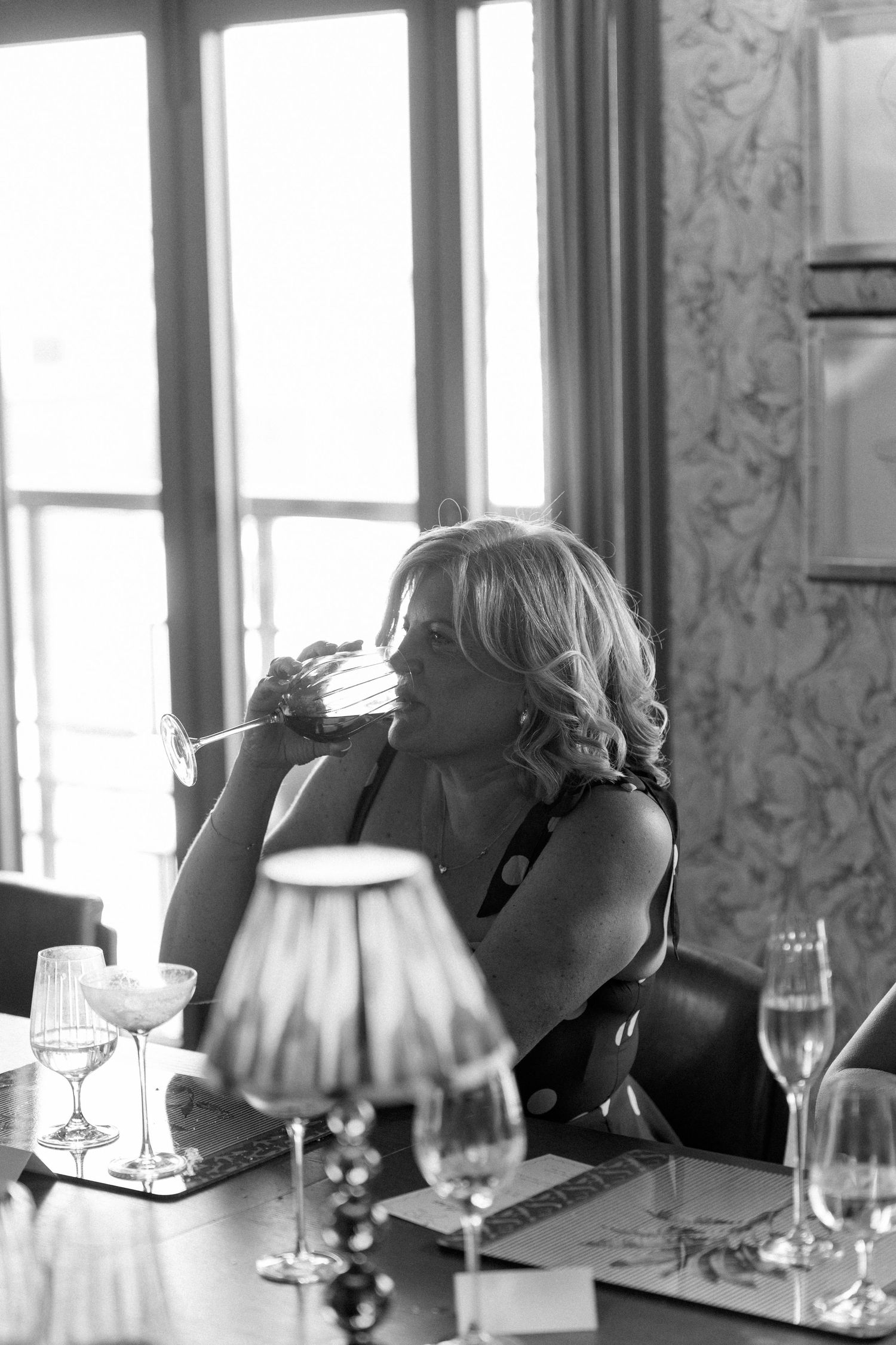 Elegant woman sips champagne at a beautifully set table near large windows in a black and white wedding photo.
