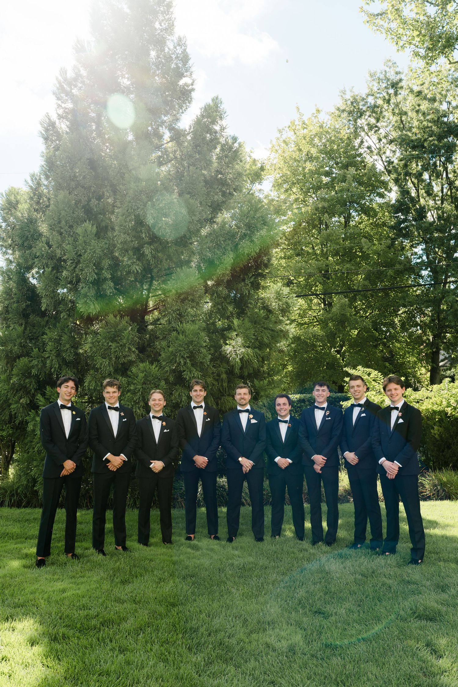 Groom and groomsmen in navy tuxedos with bow ties pose outdoors on green lawn with trees in background.