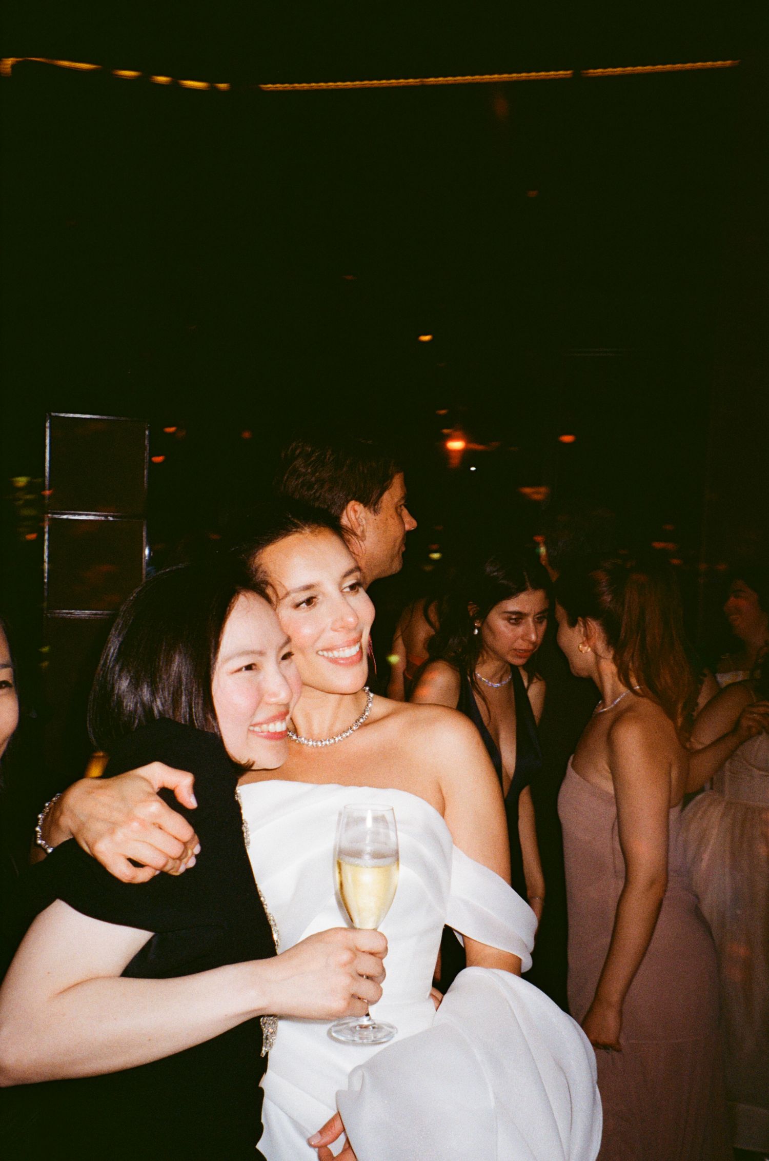 Wedding reception guests celebrating with champagne and hugs in a dimly lit venue.