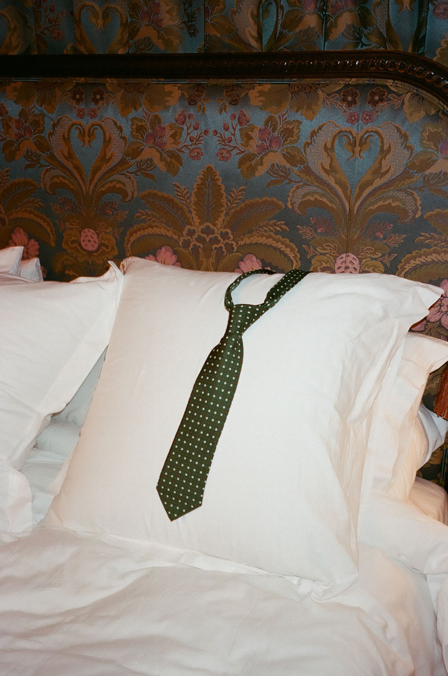 A sequence of pillows on a bed with a green polka dot necktie arranged across them against a decorative wallpaper background.