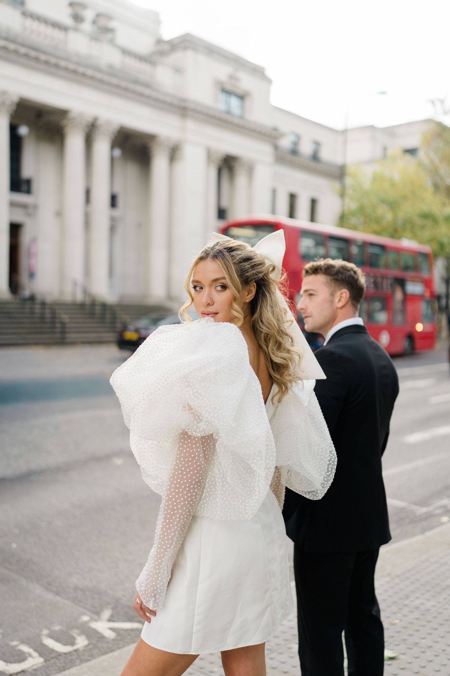 A couple in formal wear dancing and twirling on a city sidewalk near classic architecture.