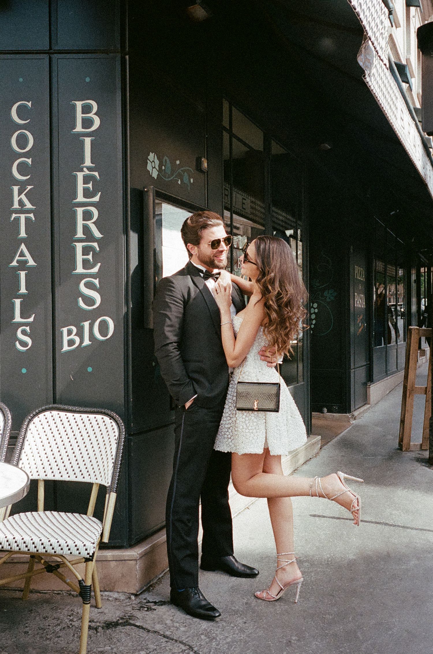 Romantic couple shares a kiss outside a French bistro cafe with classic Parisian storefront and sidewalk seating.