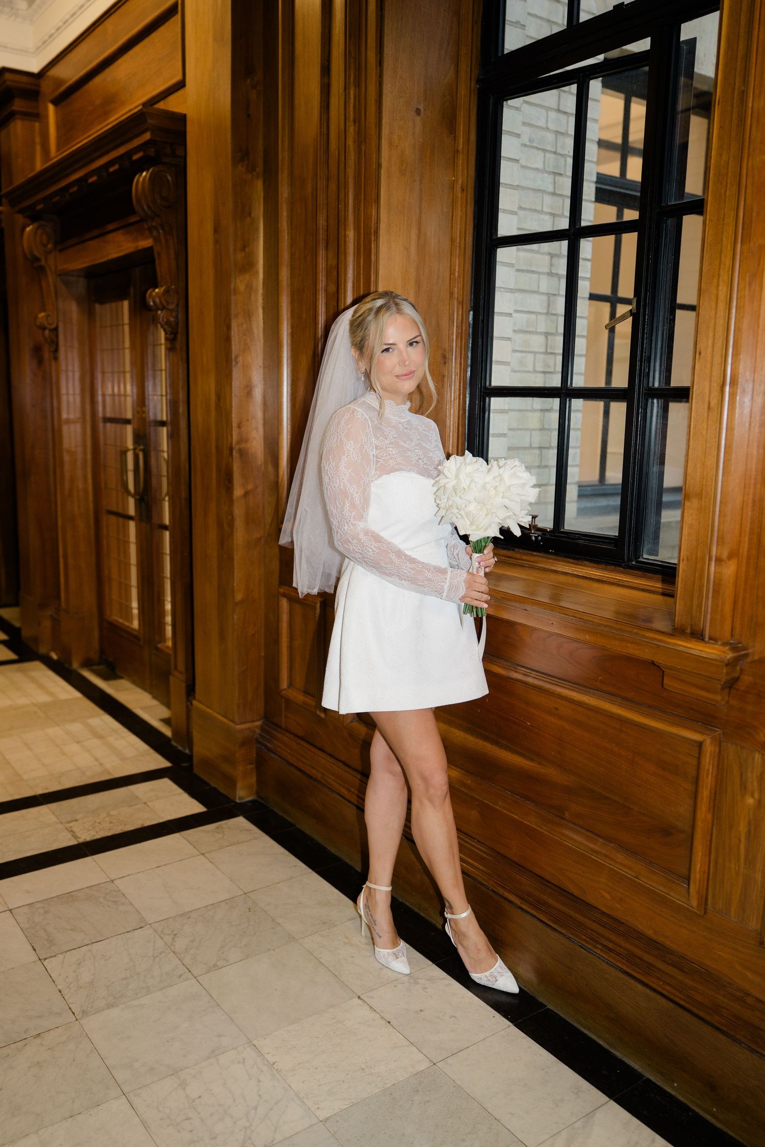 Bride in short dress walks gracefully through wood-paneled hallway.