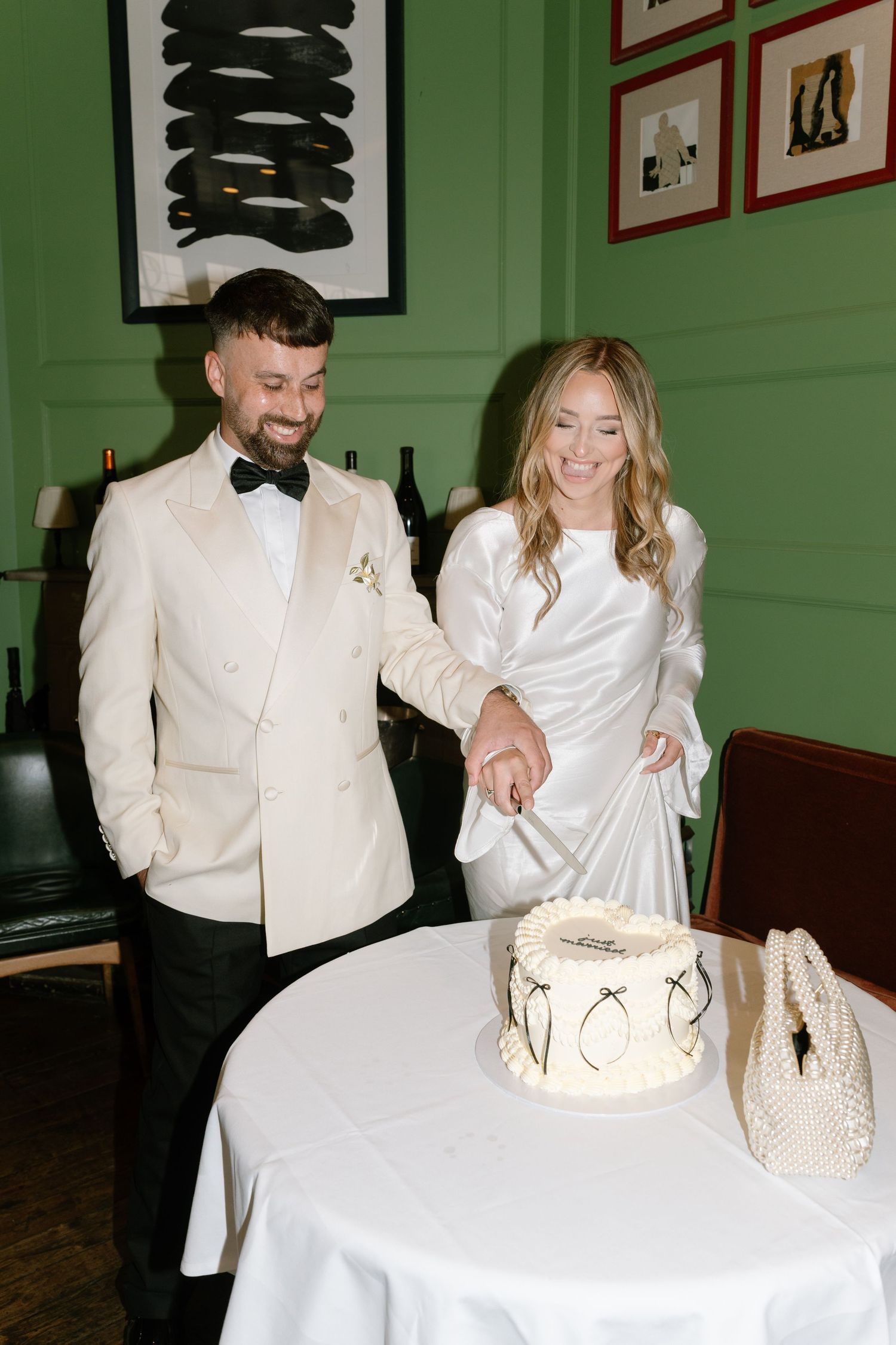Wedding couple cutting their white wedding cake in room with green walls and framed artwork.