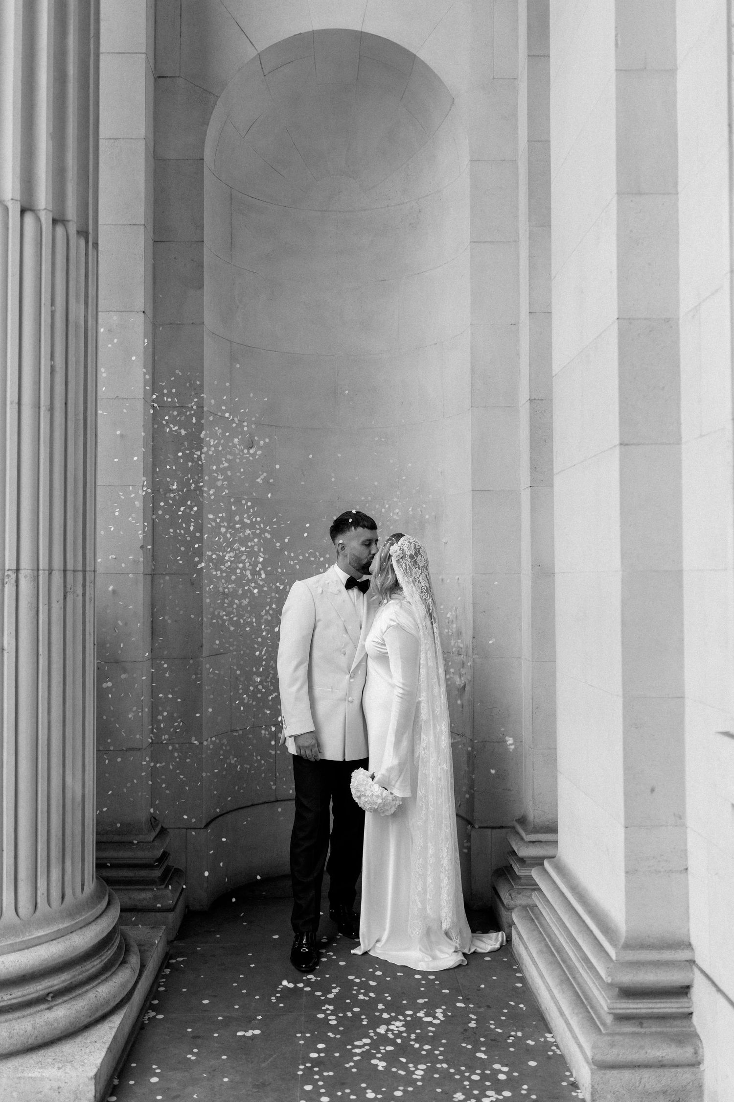 A couple shares a romantic moment among classical columns with confetti falling around them.