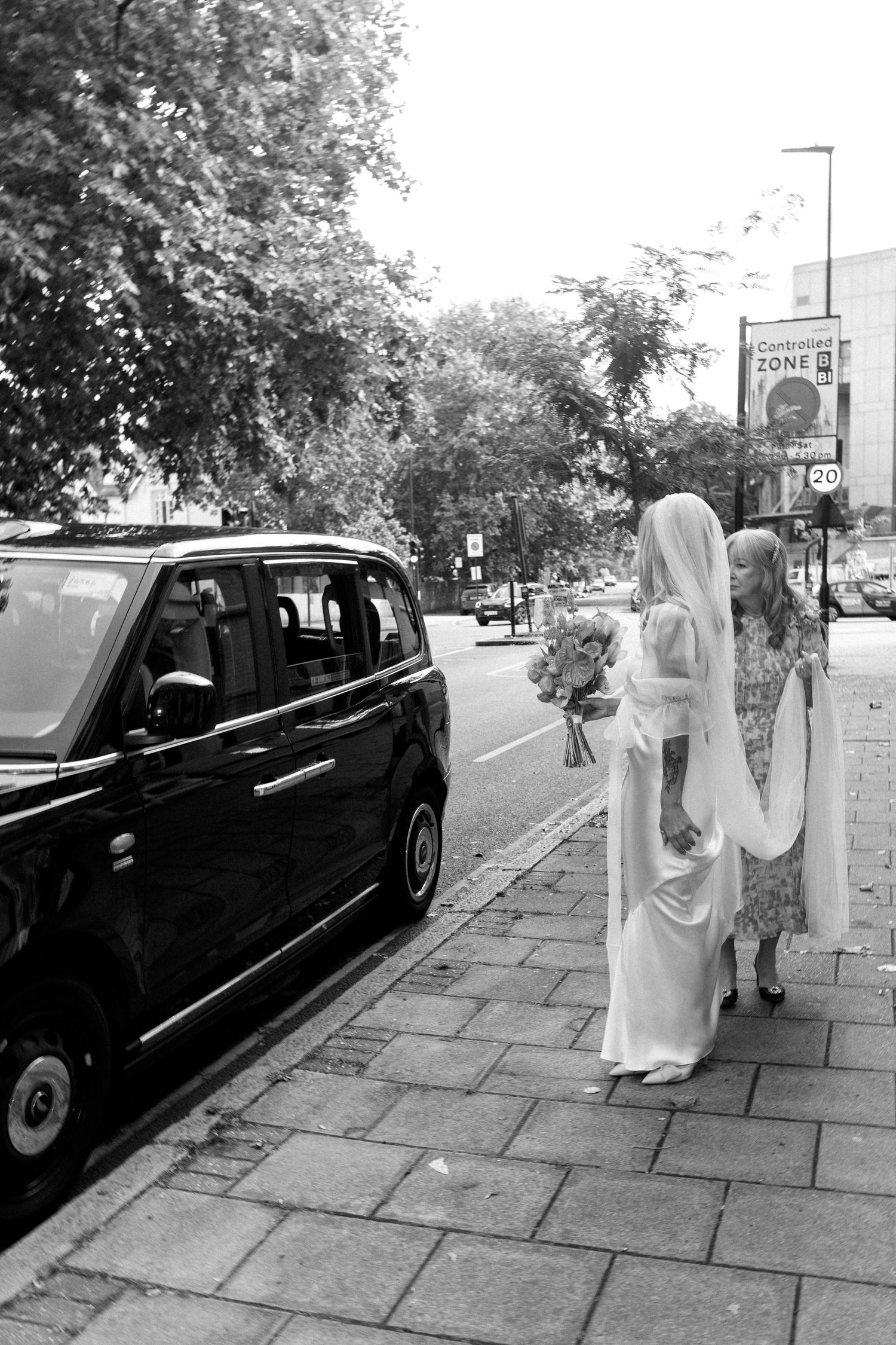 Classic black car parked on a sidewalk with figures in formal attire walking alongside it.