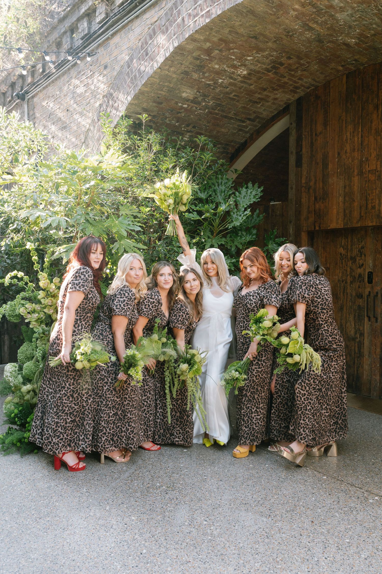 Bridesmaids in matching leopard print dresses pose playfully against a rustic brick wall backdrop.