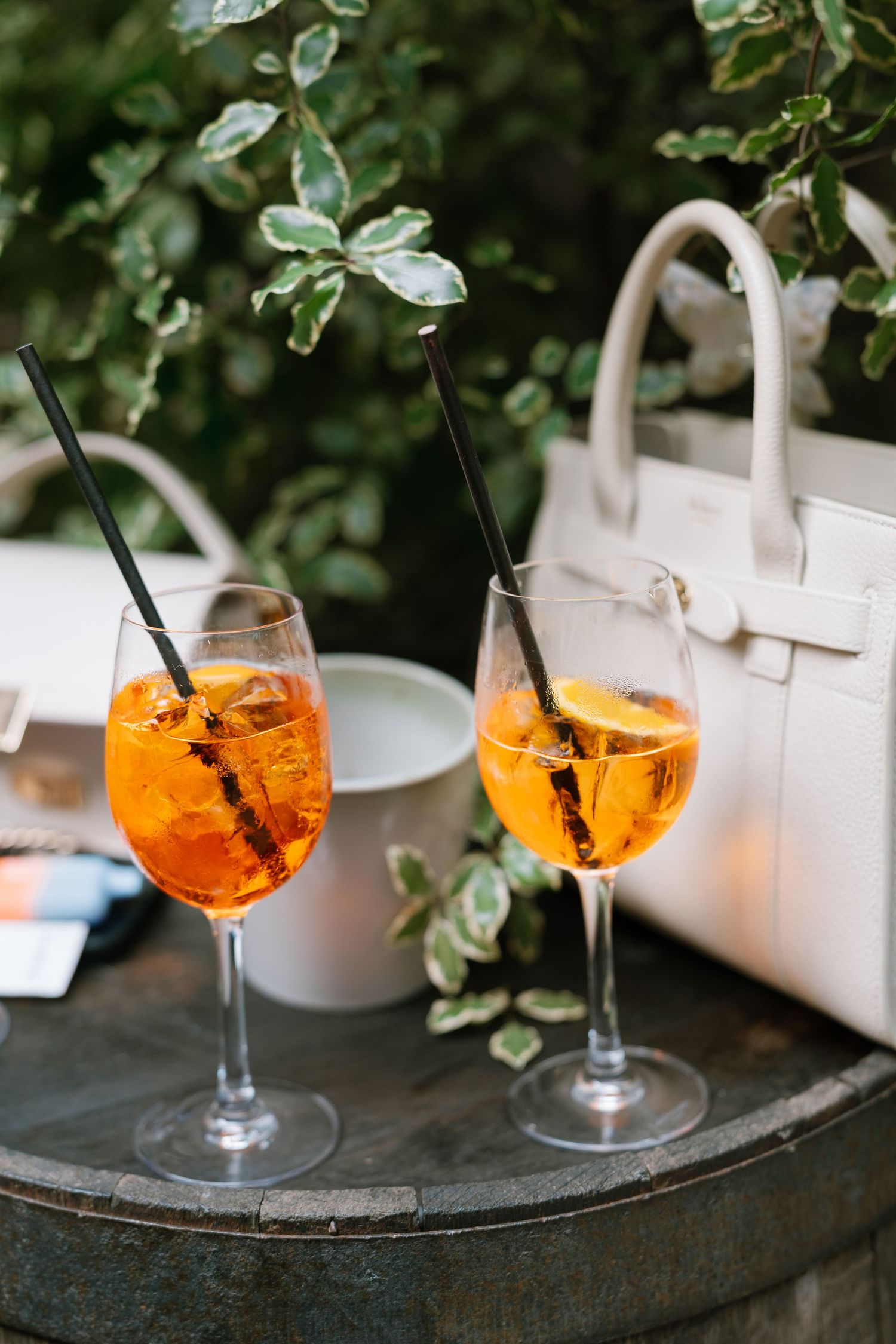 Two orange aperol spritz cocktails garnished with herbs on a dark tray with a white handbag.
