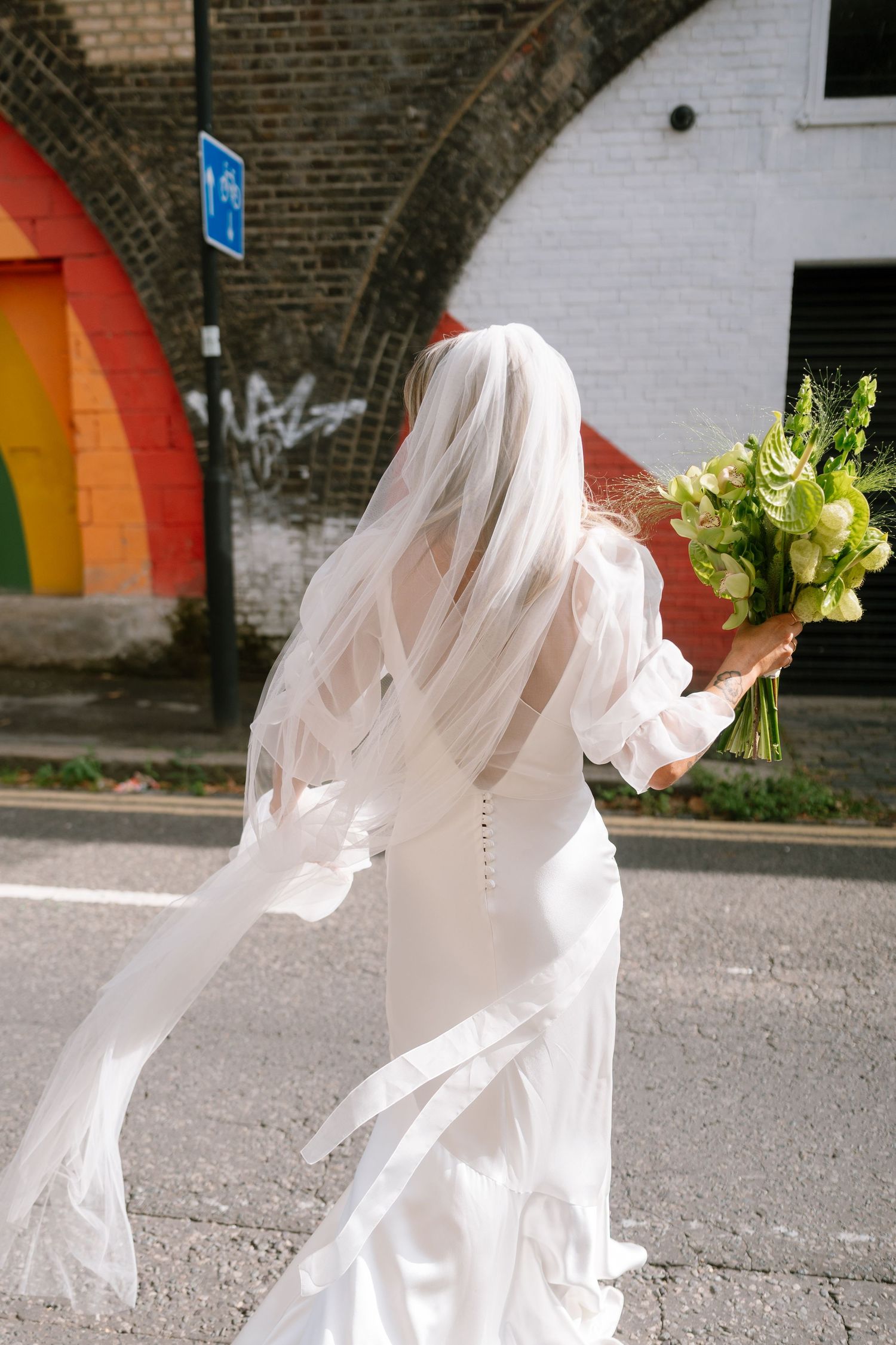 Bride in flowing white dress and veil holding bouquet against colorful wall.