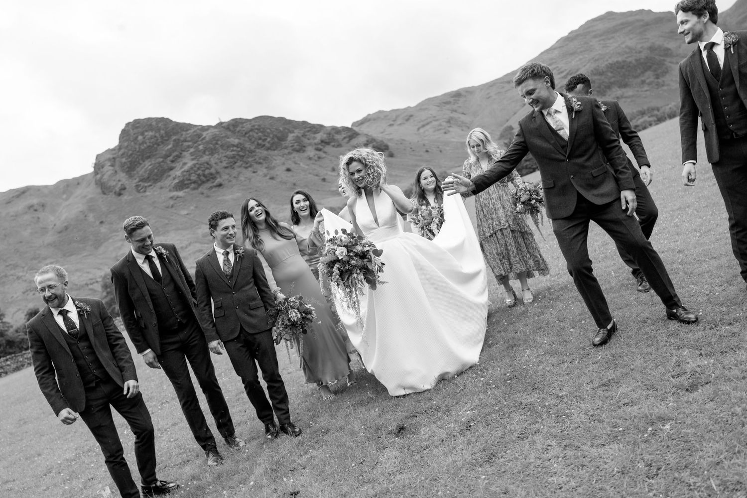 Wedding party walks across a grassy hillside with mountains in the background.