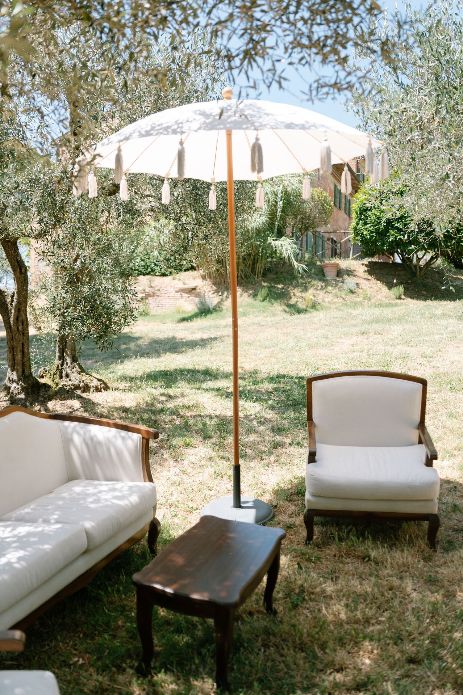 White lounge chairs and umbrellas arranged outdoors under olive trees creating a relaxing seating area.