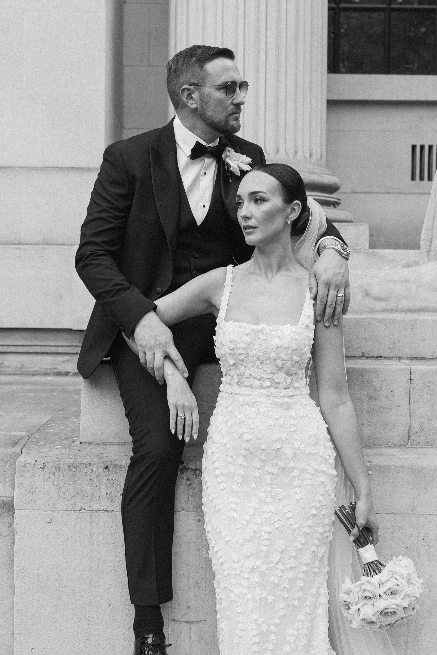 Stylish wedding couple poses against white stone columns of historic building.