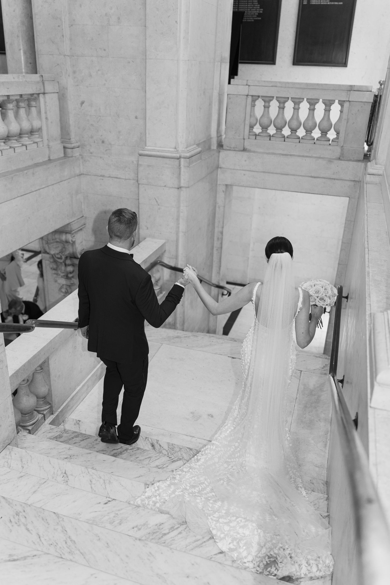 Black and white photo of newlyweds walking down marble stairs with ornate railings.