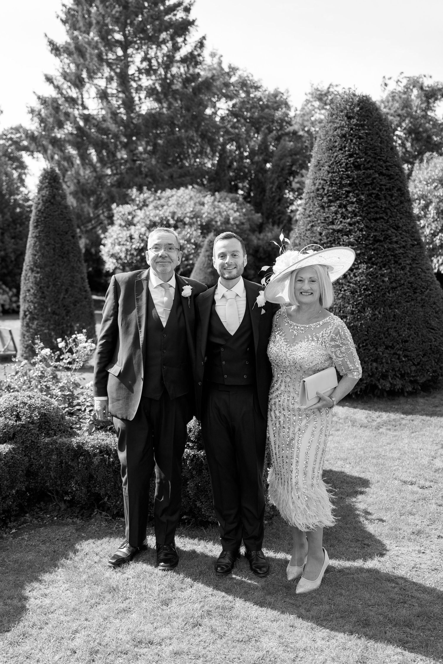 Wedding party members stand together on manicured grounds for formal black and white photographs.