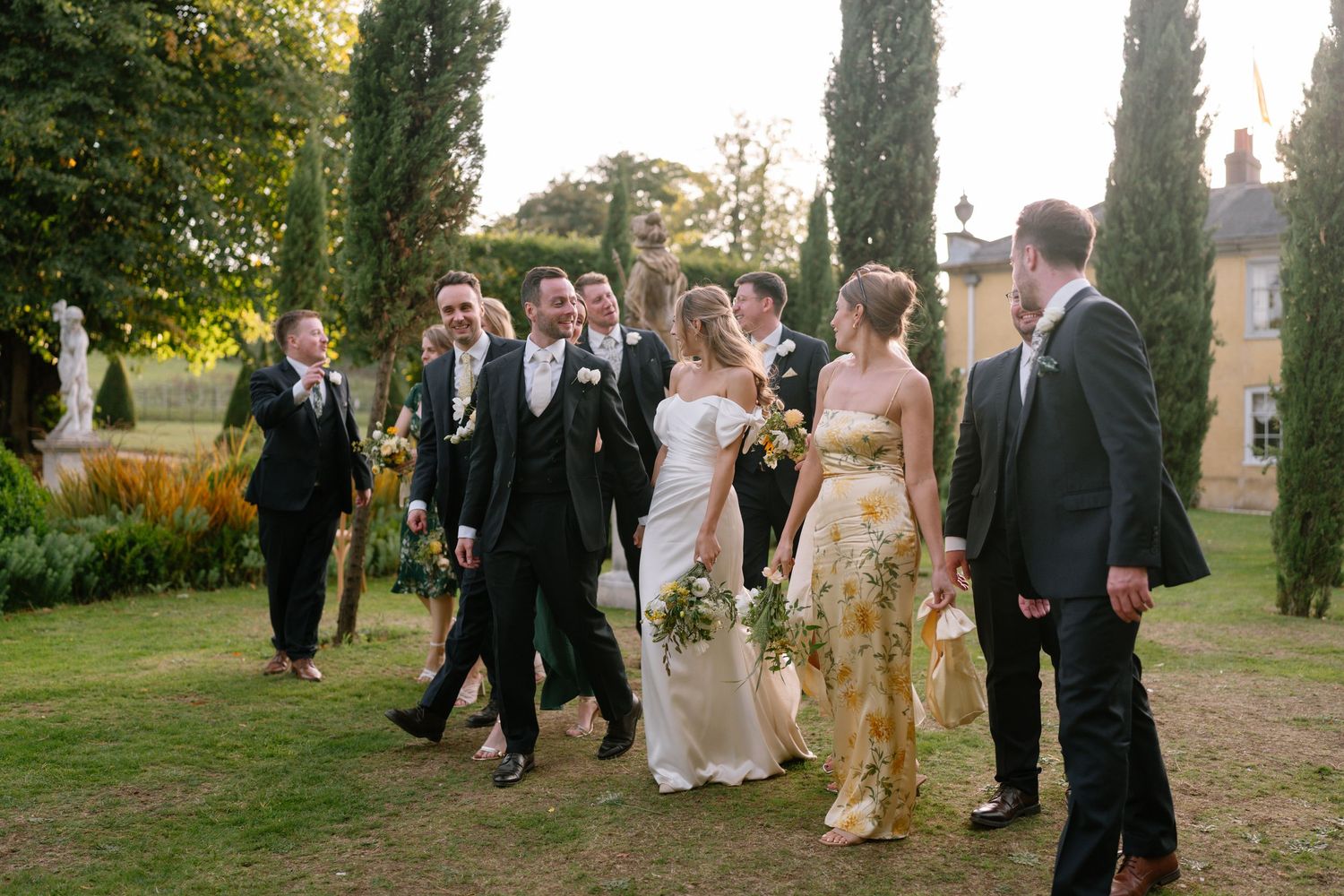 Wedding party mingles on lawn during golden hour with cypress trees creating vertical lines.