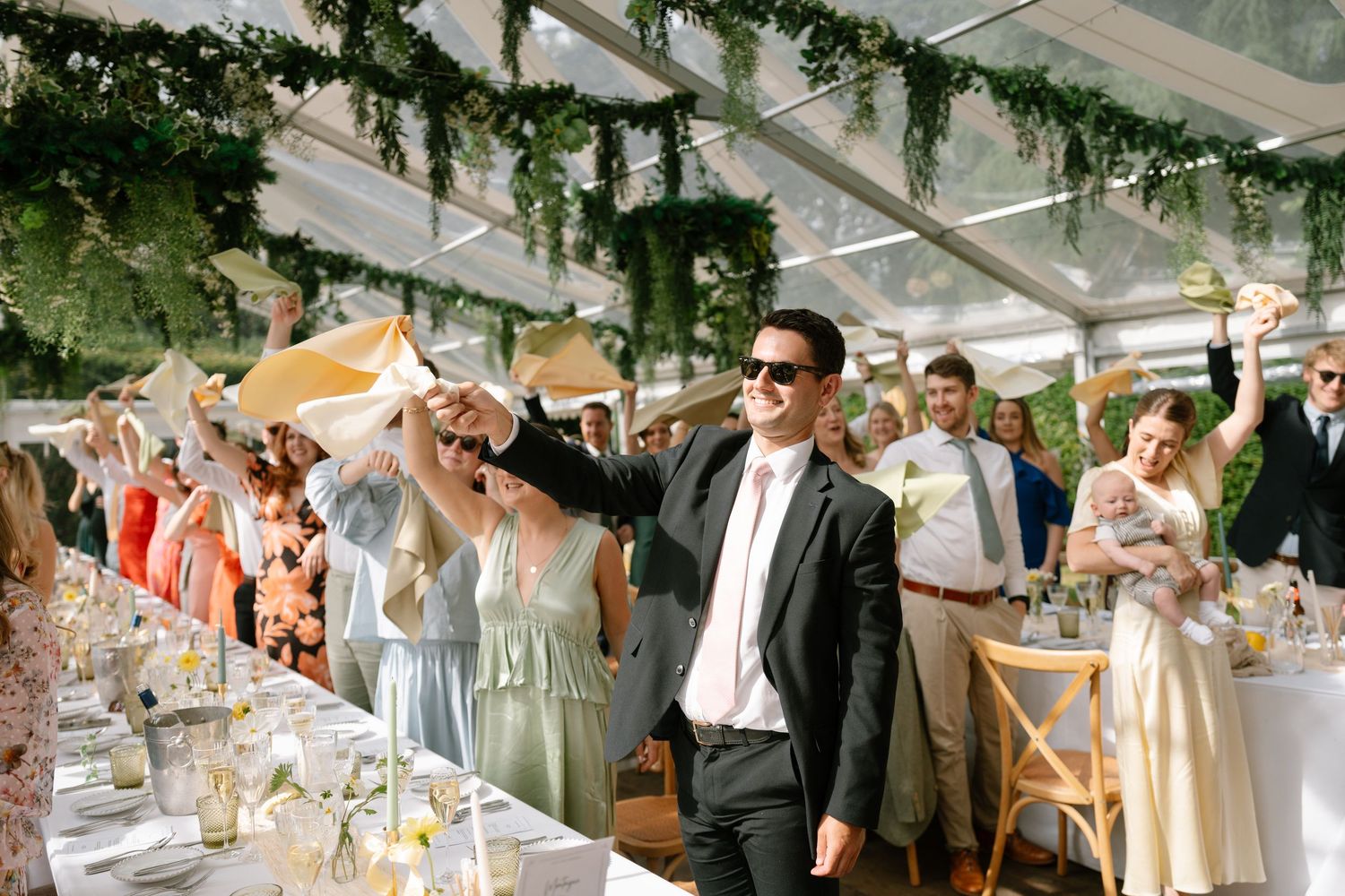 Wedding guests celebrating with napkin wave during outdoor reception under tent.