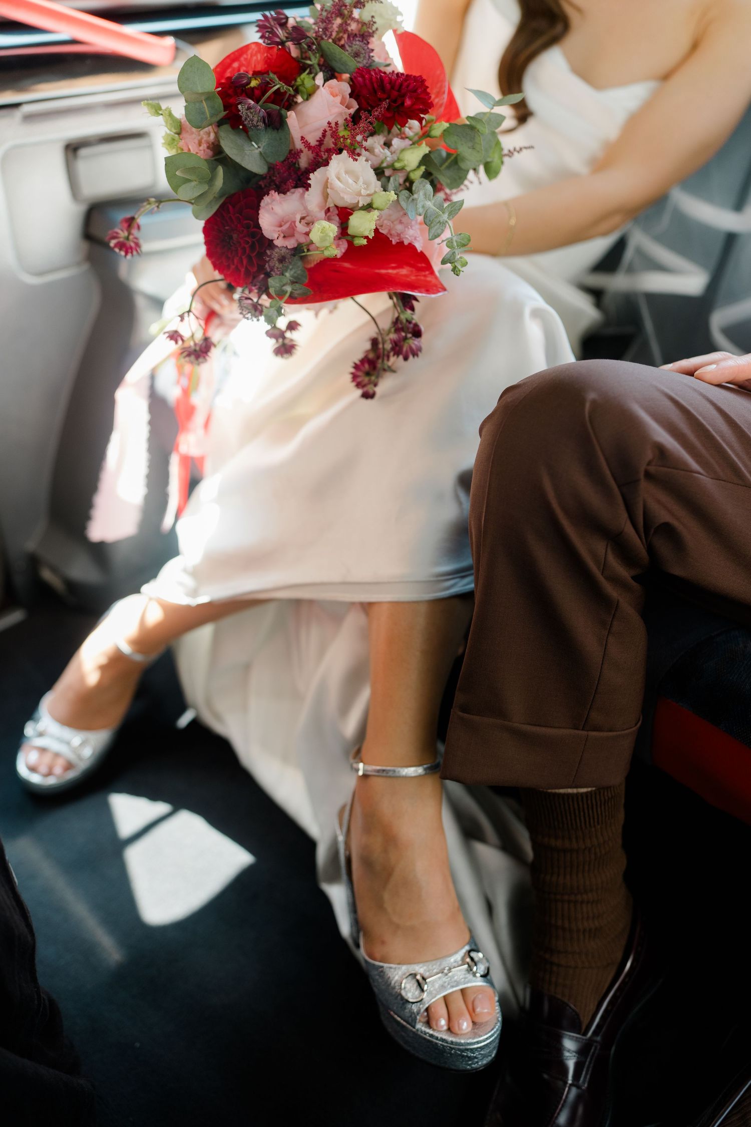 Bride in white satin dress and silver heels sits beside groom in brown suit holding a vibrant red floral bouquet.